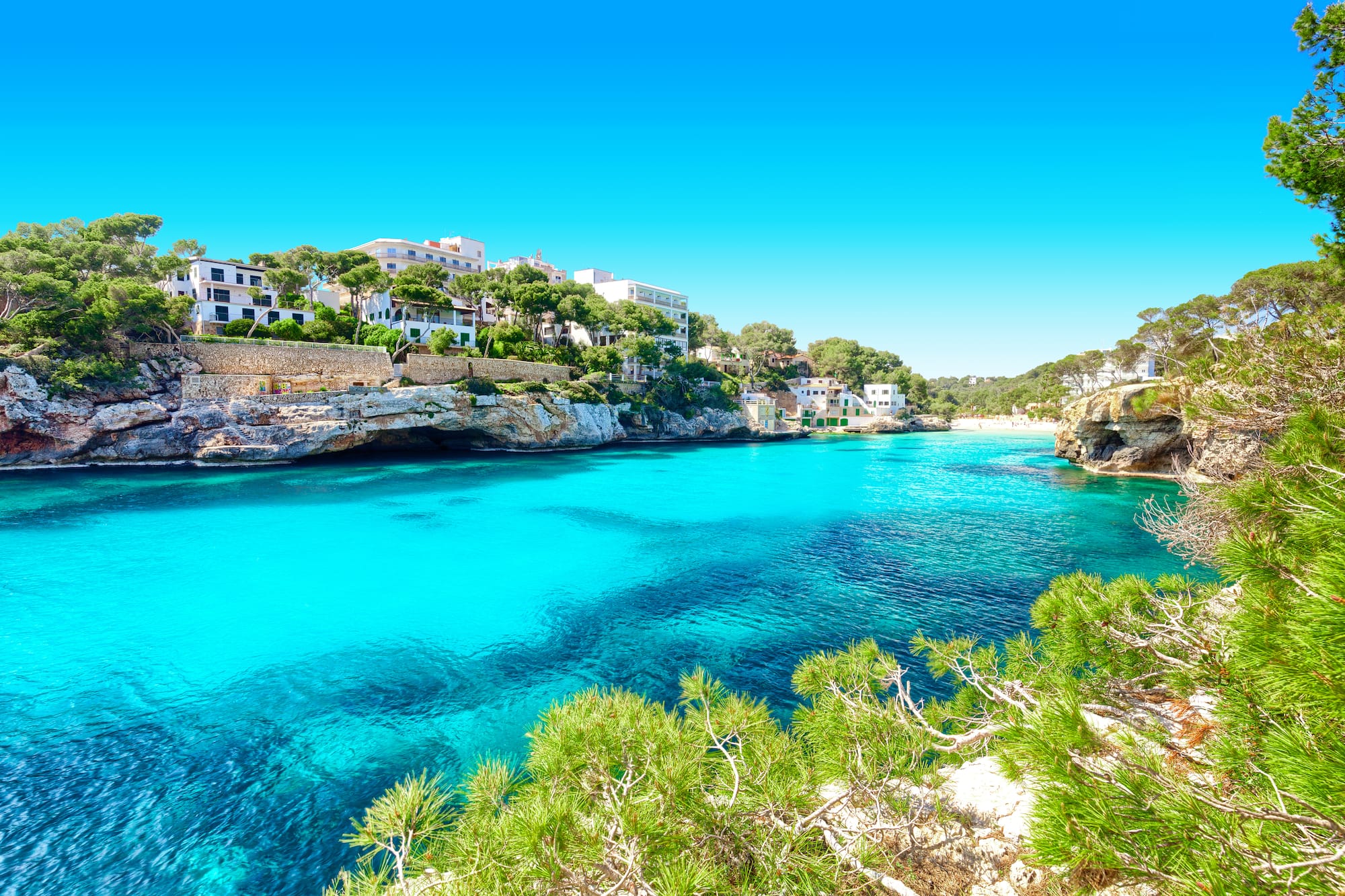 Cala con aguas turquesas, acantilados rocosos y casas rodeadas de pinos.