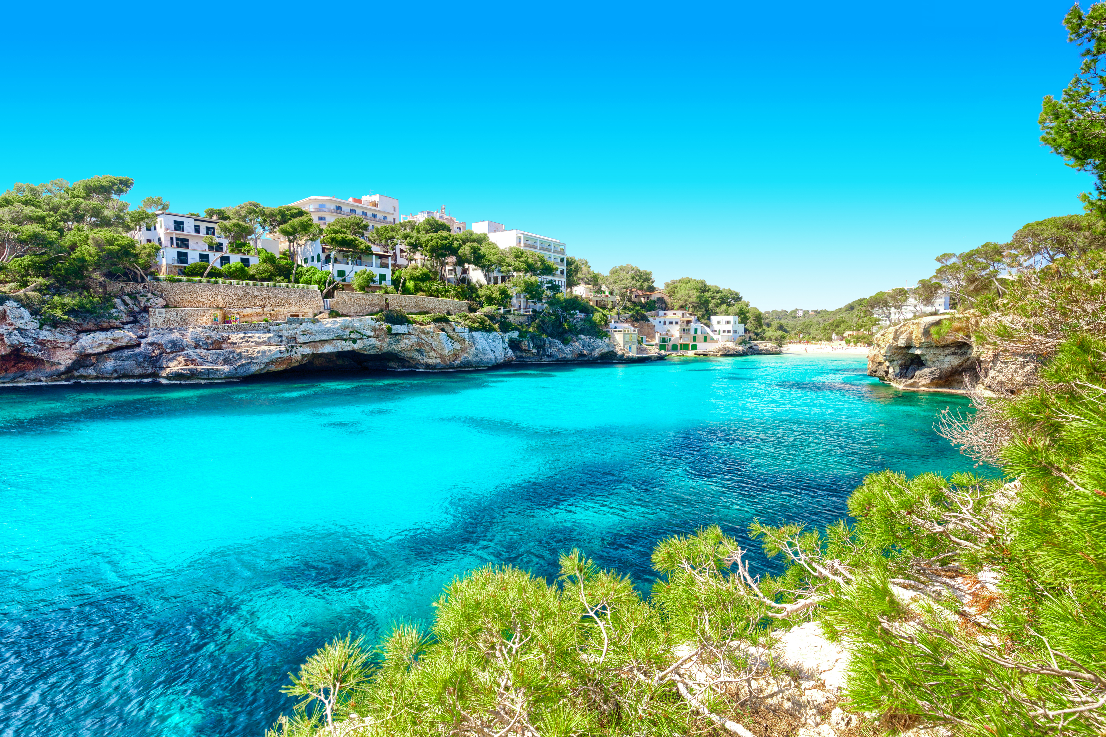Cala con aguas turquesas, acantilados rocosos y casas rodeadas de pinos.