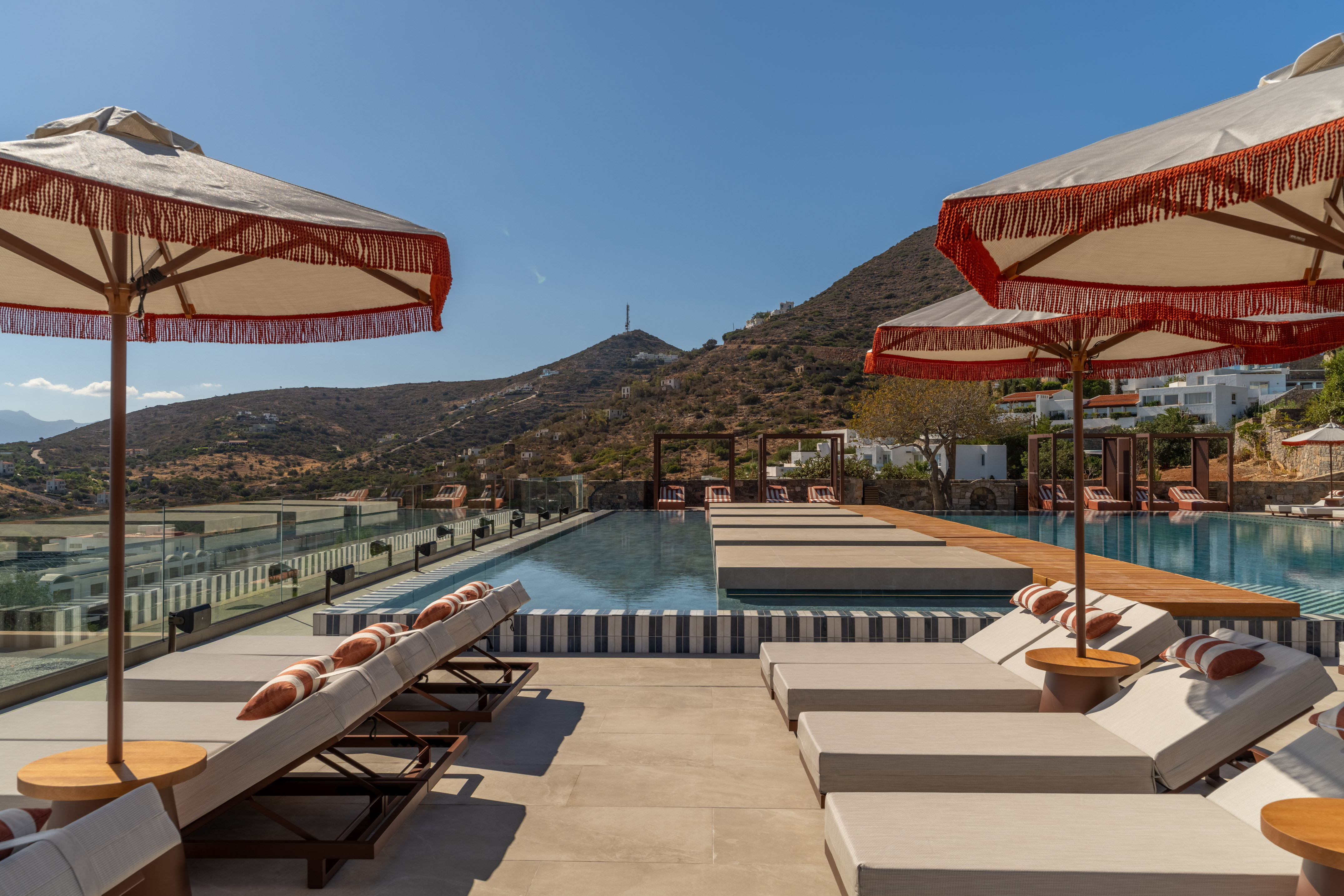 Resort infinity pool deck with sun loungers, fringed umbrellas, mountains.