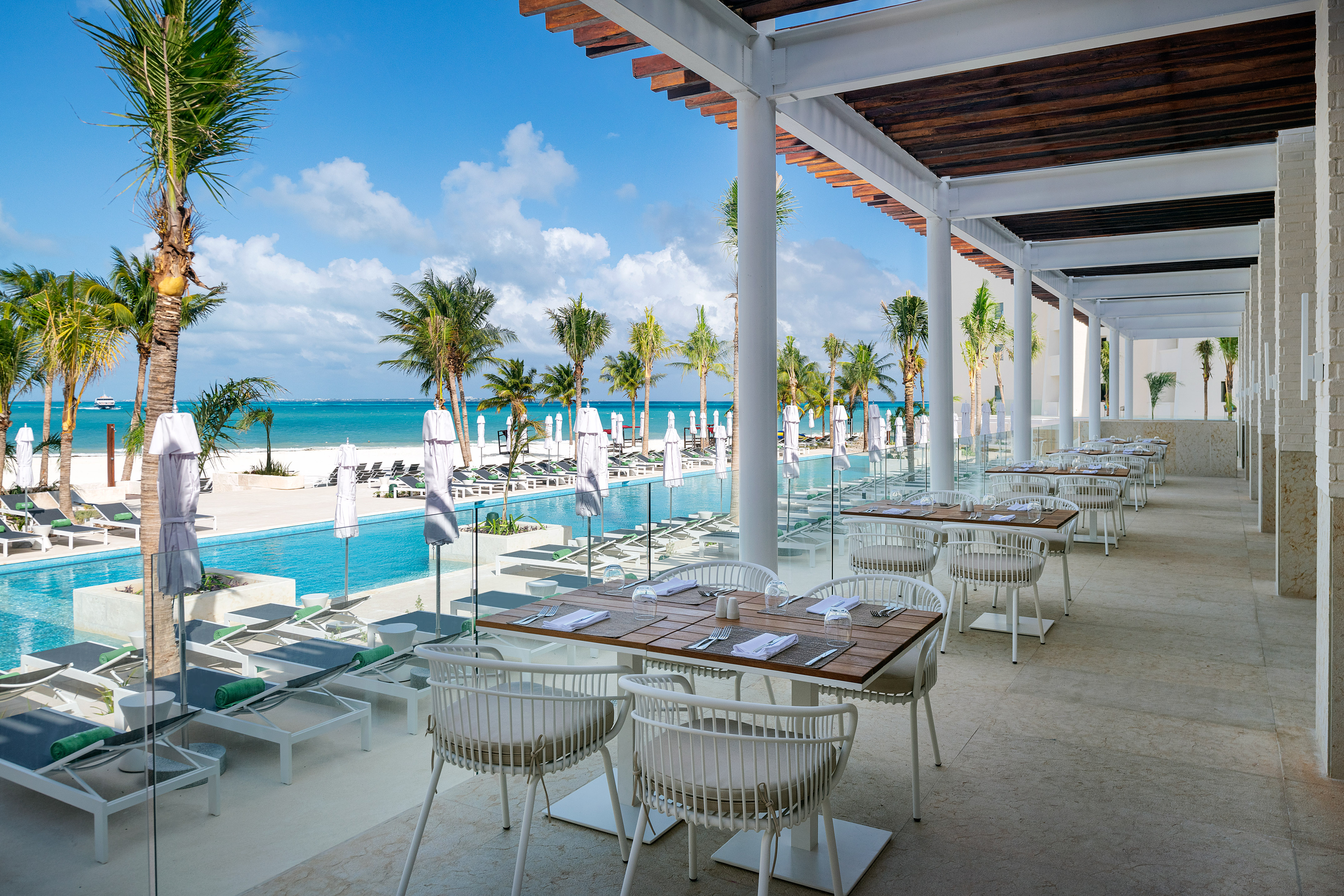 Beachfront restaurant with outdoor tables, pool, palm trees, and ocean view.