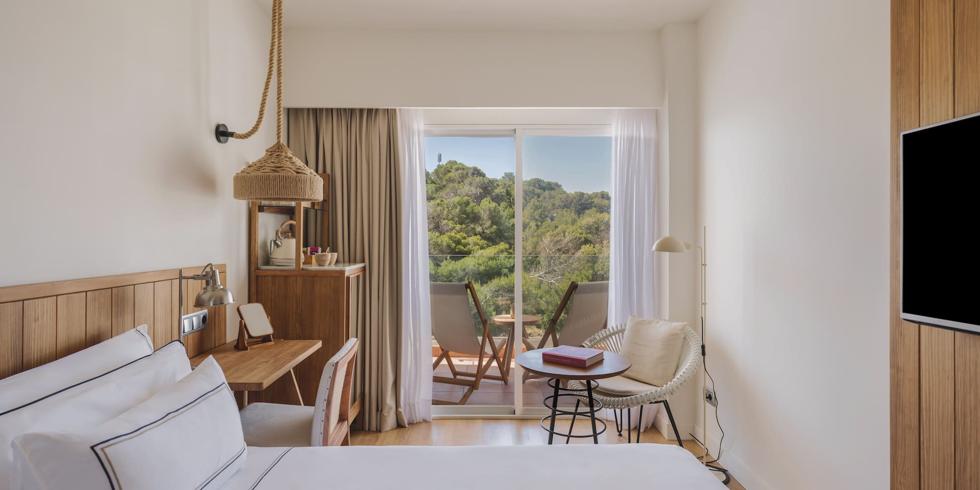 Modern hotel room with white bed, wooden desk, and balcony forest view.