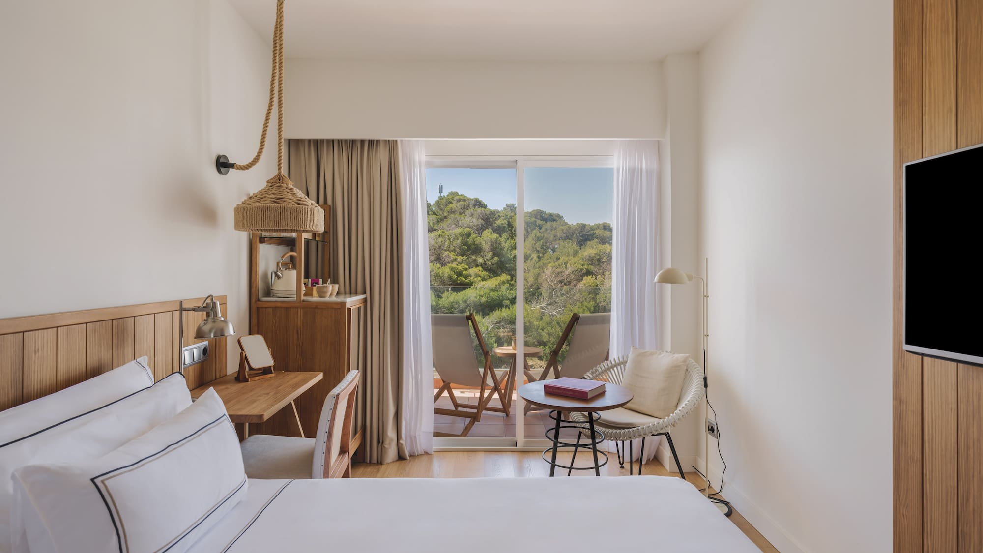 Modern hotel room with white bed, wooden desk, and balcony forest view.