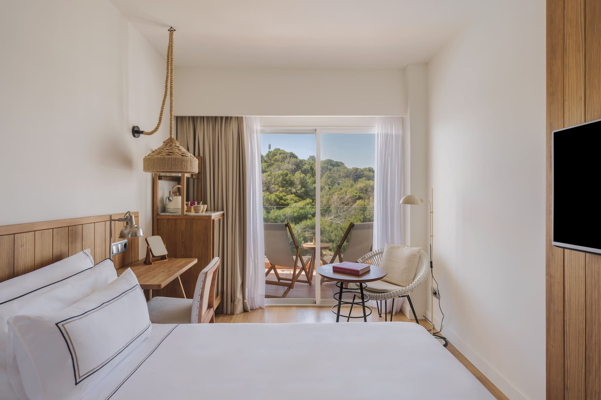 Modern hotel room with white bed, wooden desk, and balcony forest view.