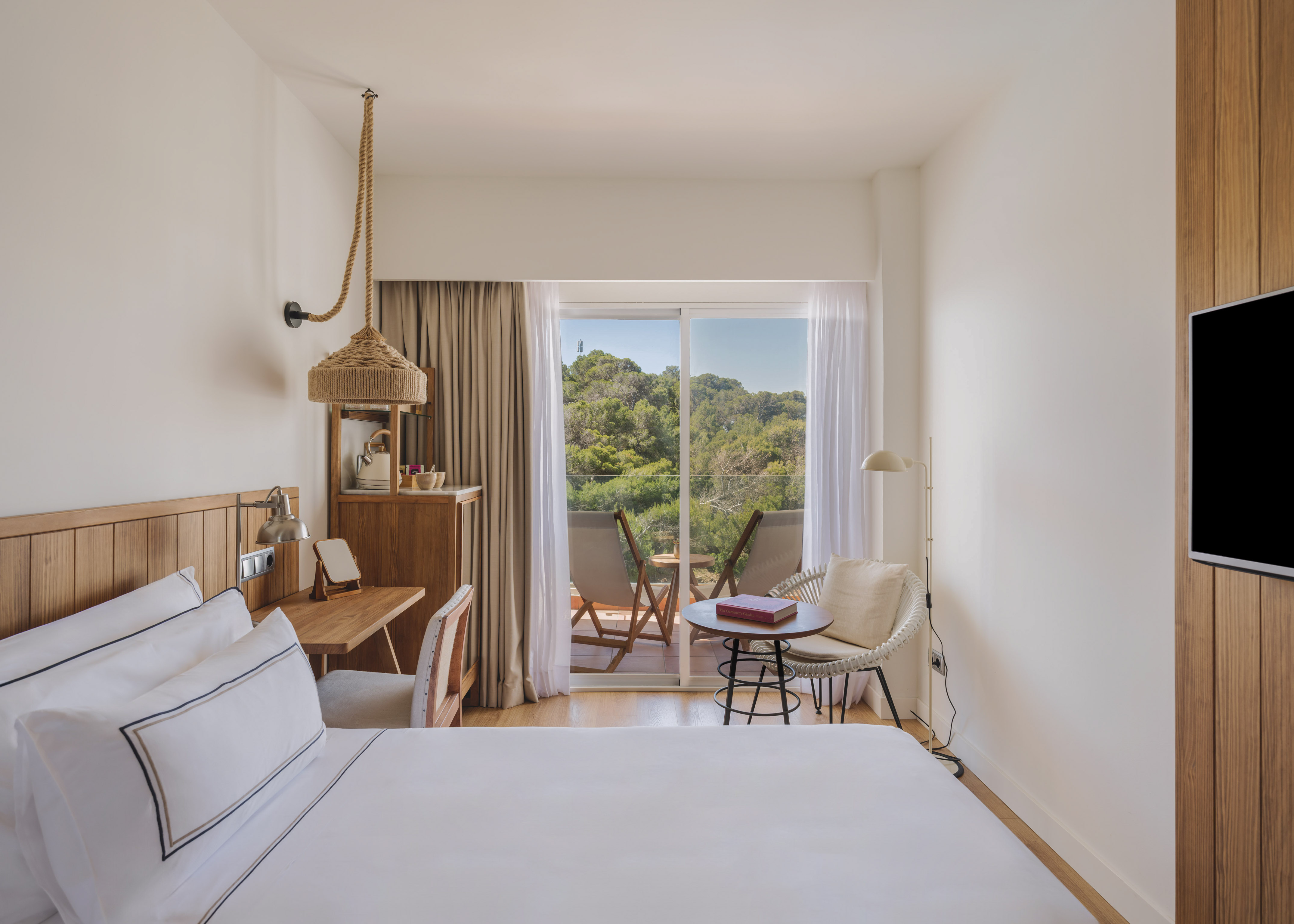 Modern hotel room with white bed, wooden desk, and balcony forest view.