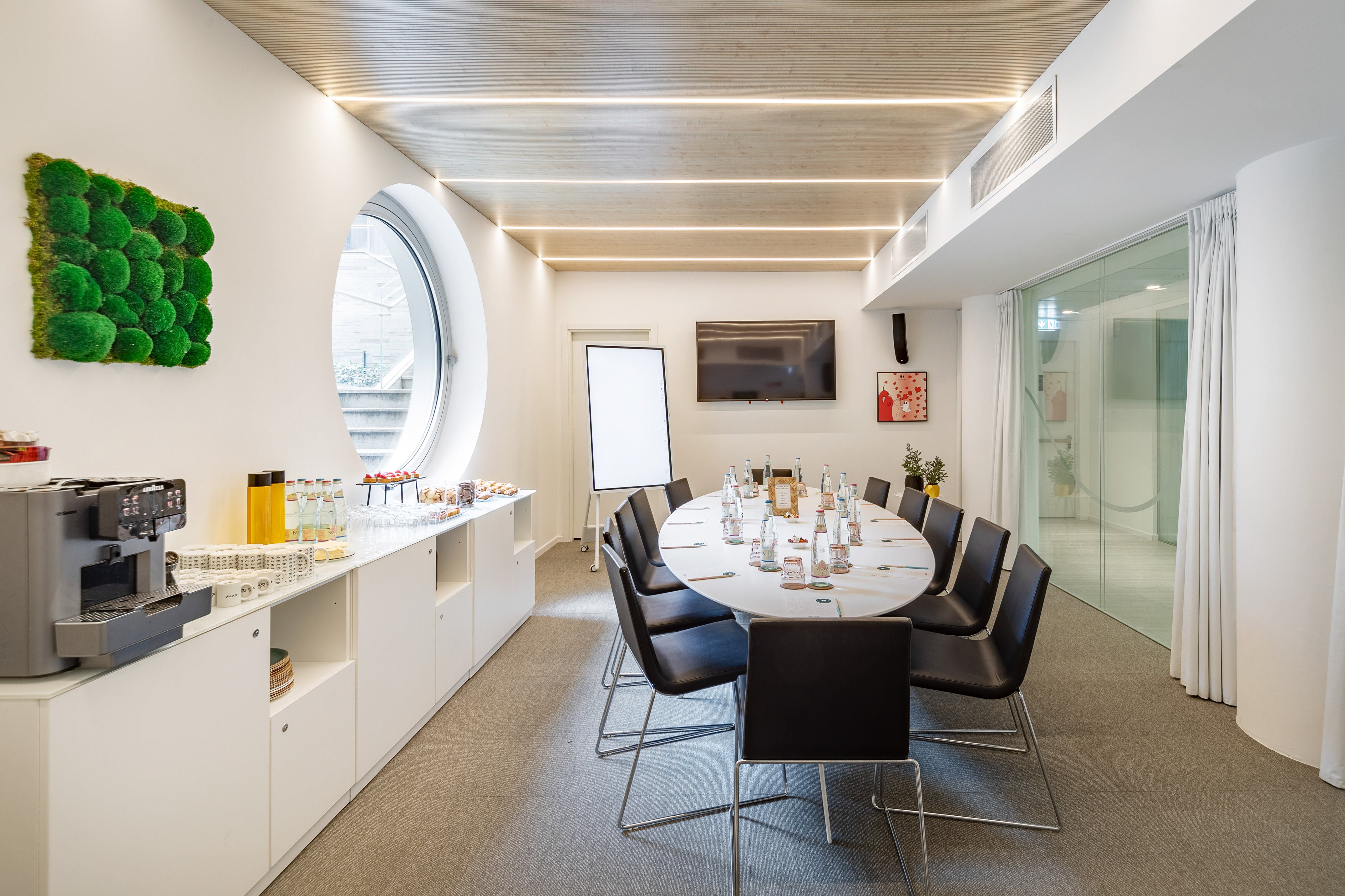 Modern conference room with oval table, TV, whiteboard, and refreshment buffet.