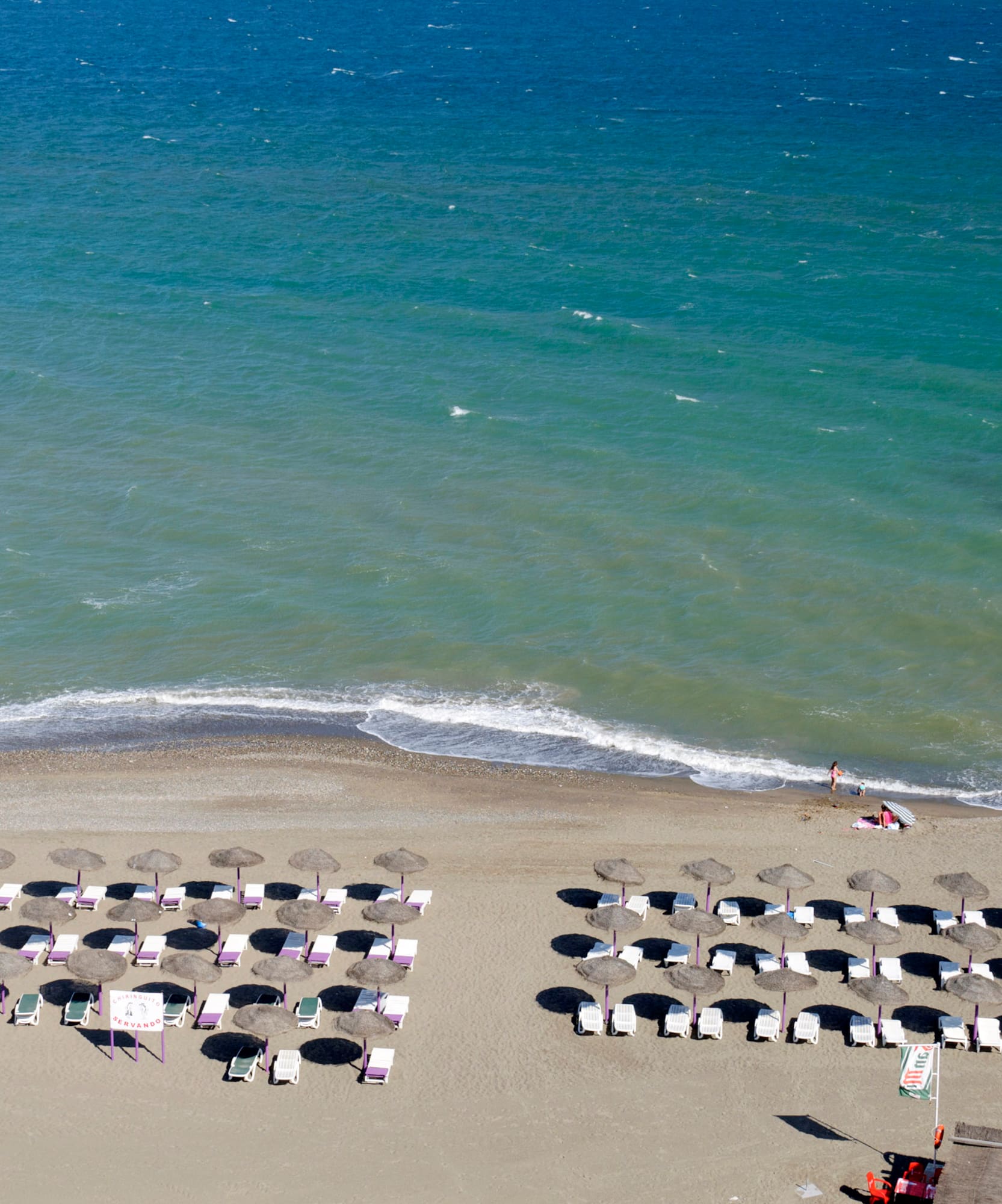 Vista aérea de playa con arena, sombrillas, tumbonas y mar turquesa.