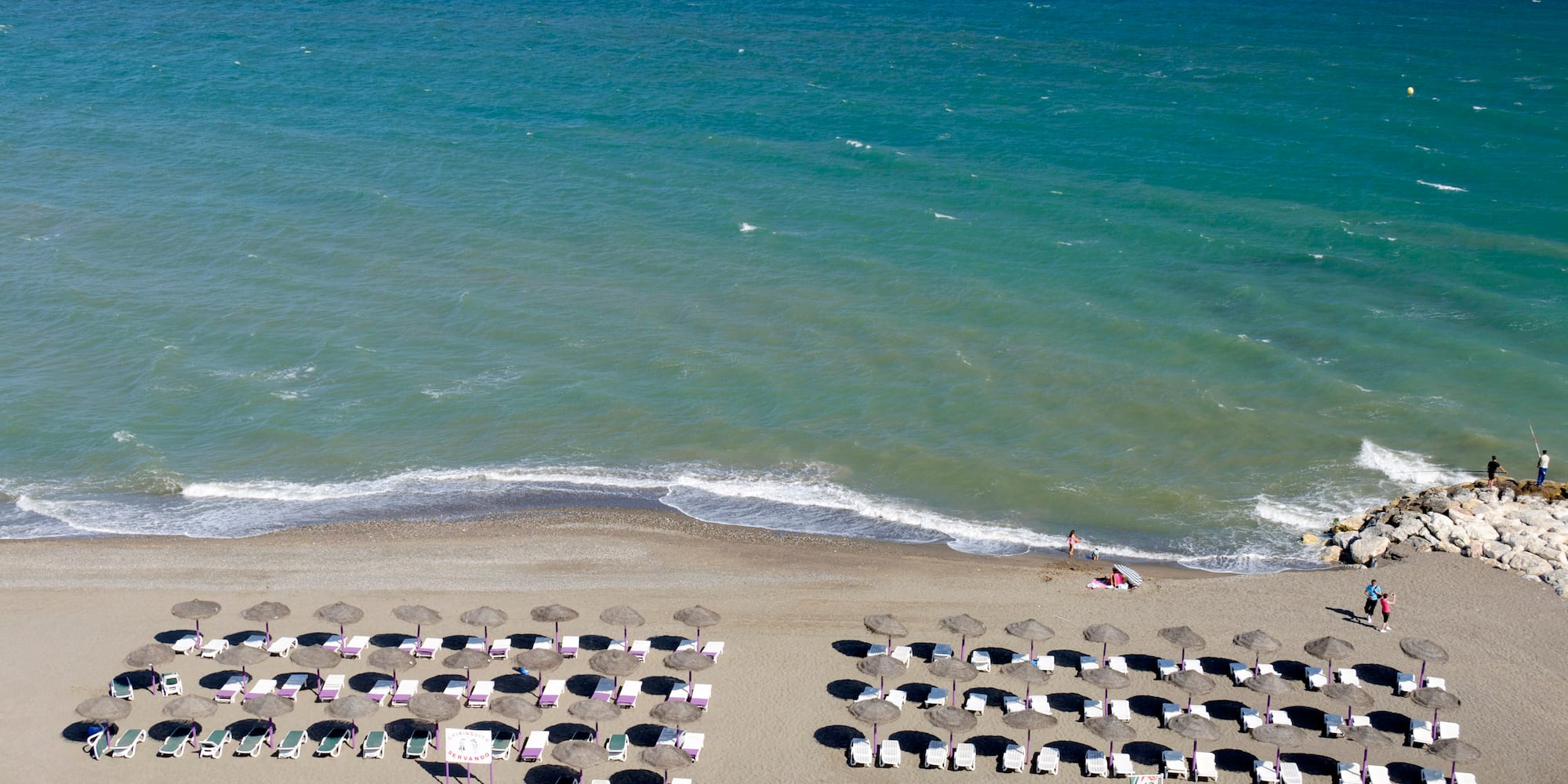 Vista aérea de playa soleada con mar turquesa, sombrillas y tumbonas en la arena.