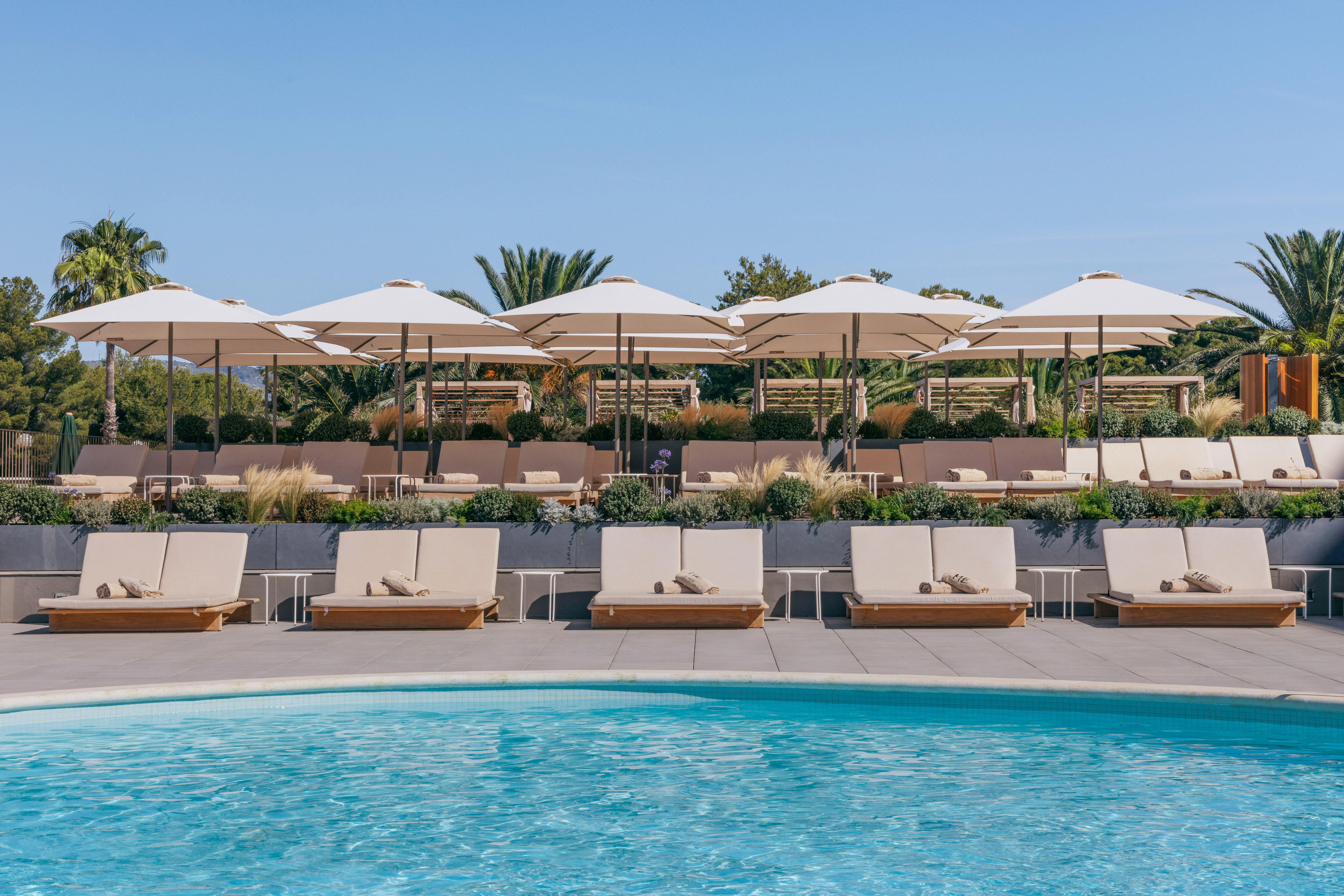 Hotel poolside with beige daybeds, white umbrellas, and tropical plants.