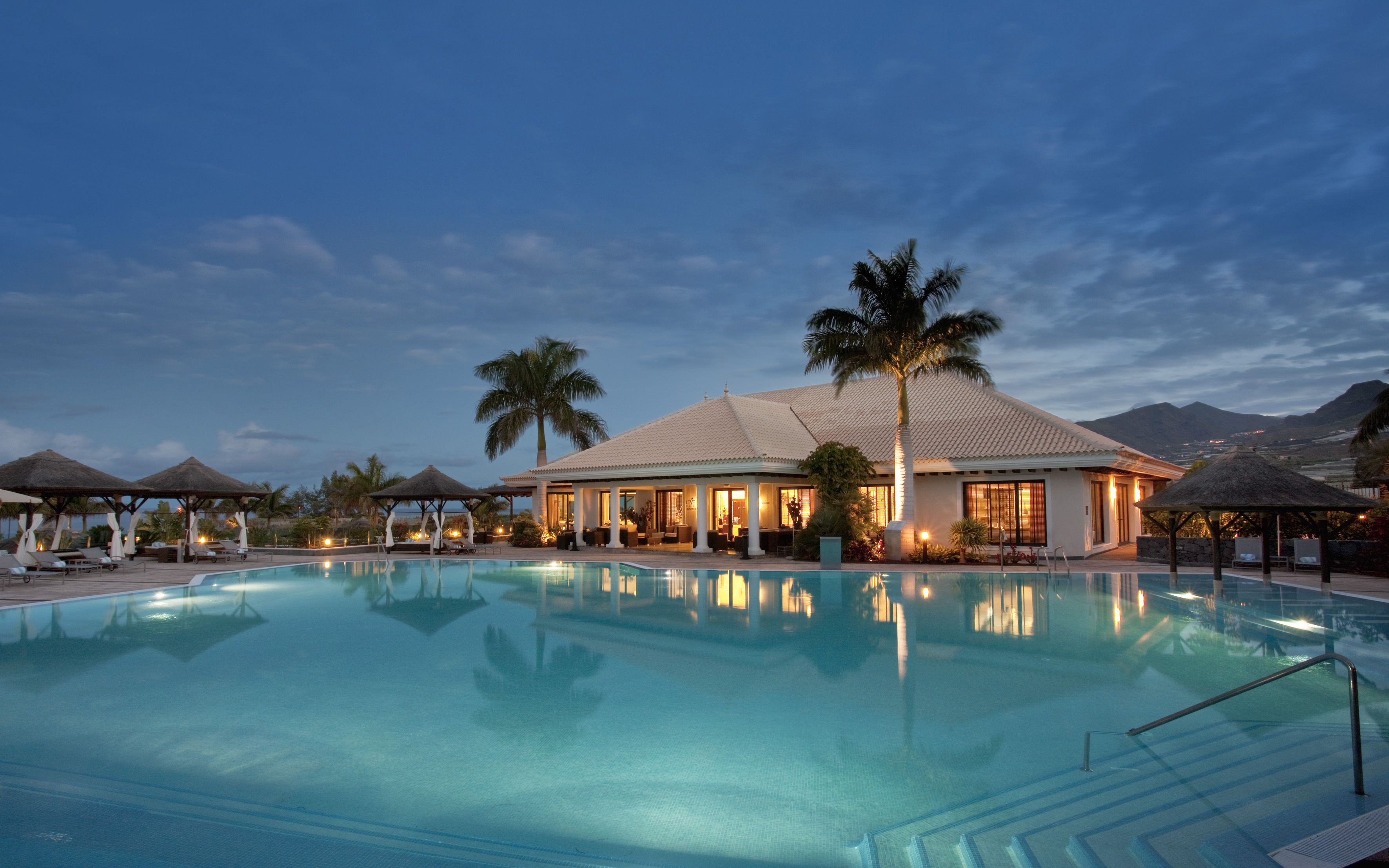 Tropical resort pool with lit building, palm trees, and mountains at twilight.