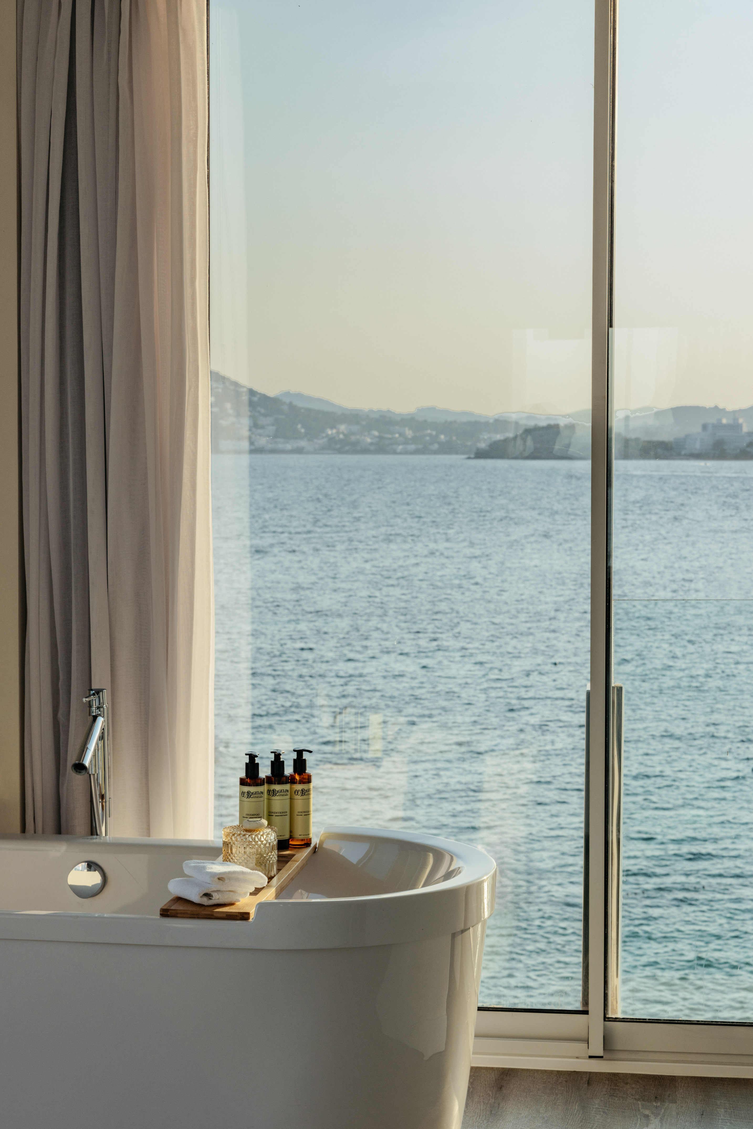 Bathtub with amenities by large window, expansive ocean view and distant mountains.