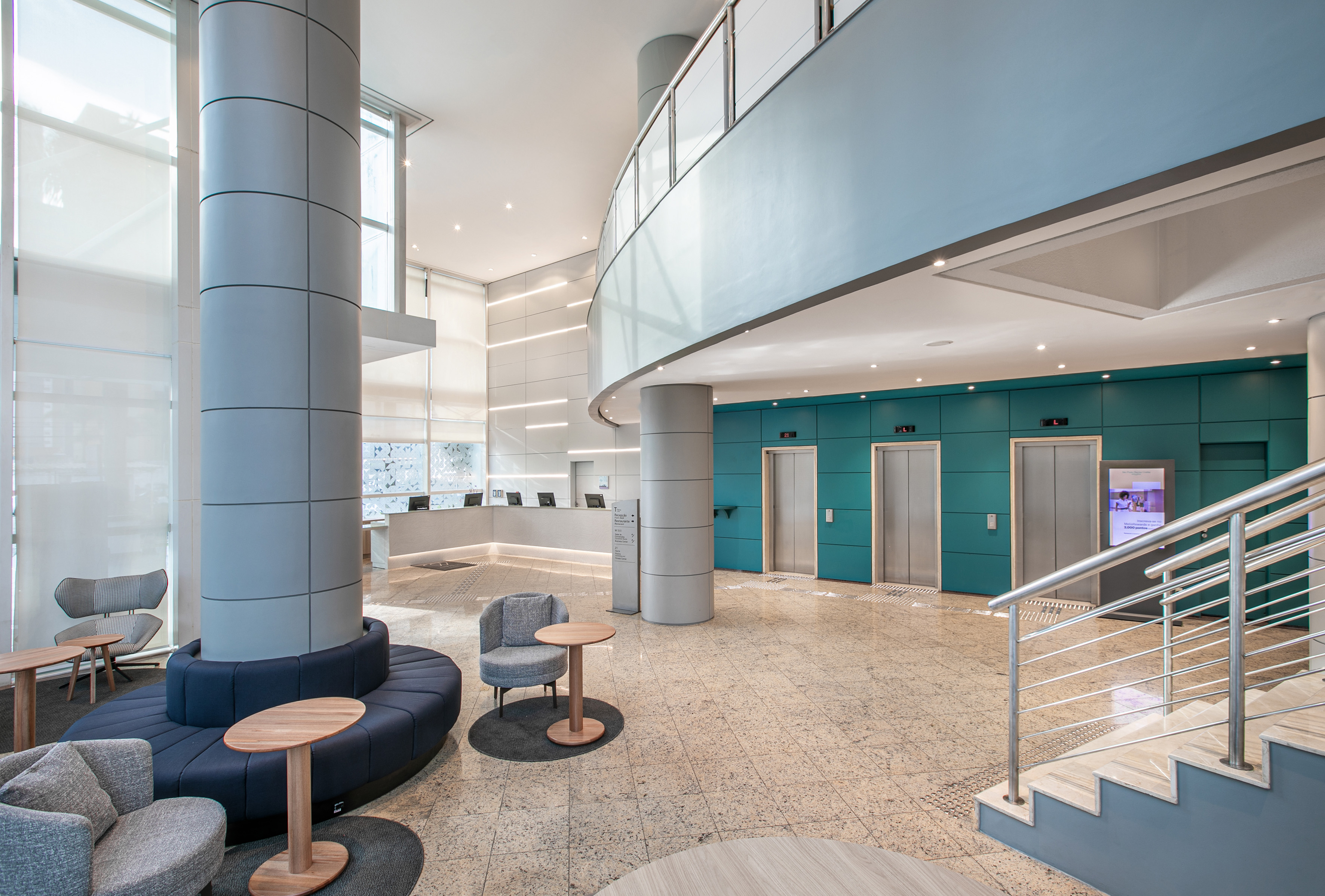 Modern hotel lobby with reception, blue seating, teal elevators, and curved mezzanine.
