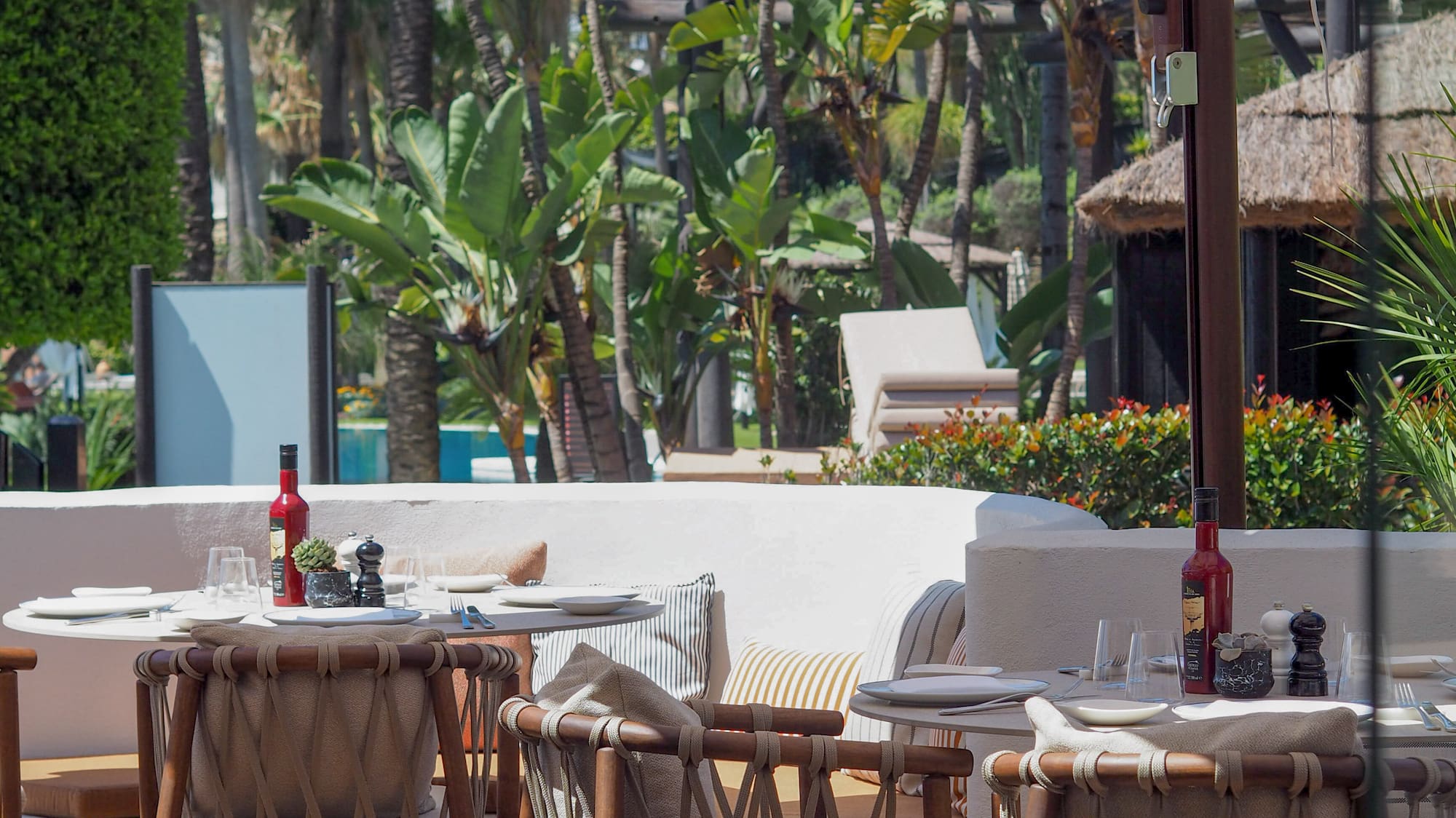 Shaded restaurant patio with wicker chairs, tables, and tropical greenery.