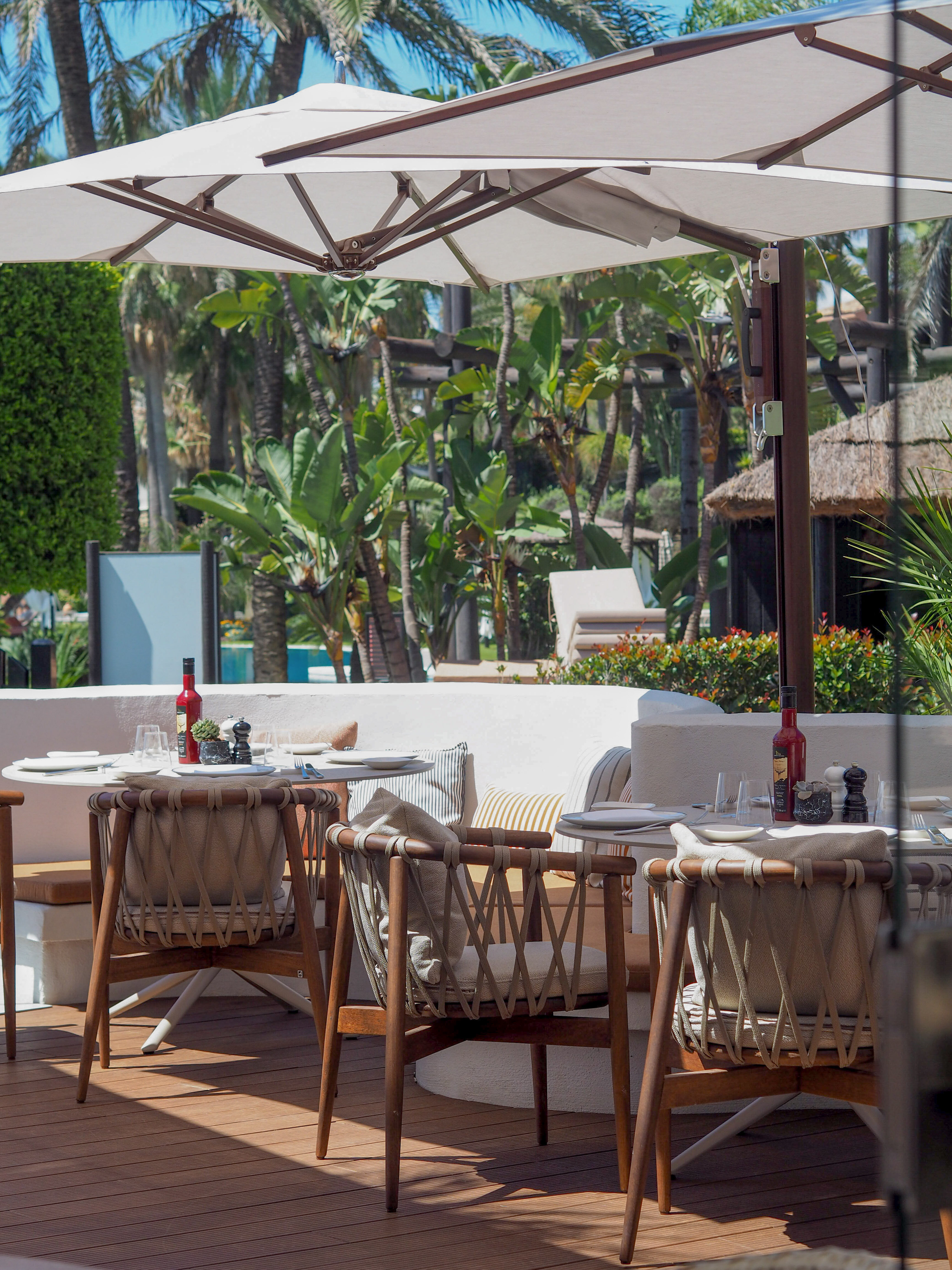 Shaded restaurant patio with wicker chairs, tables, and tropical greenery.