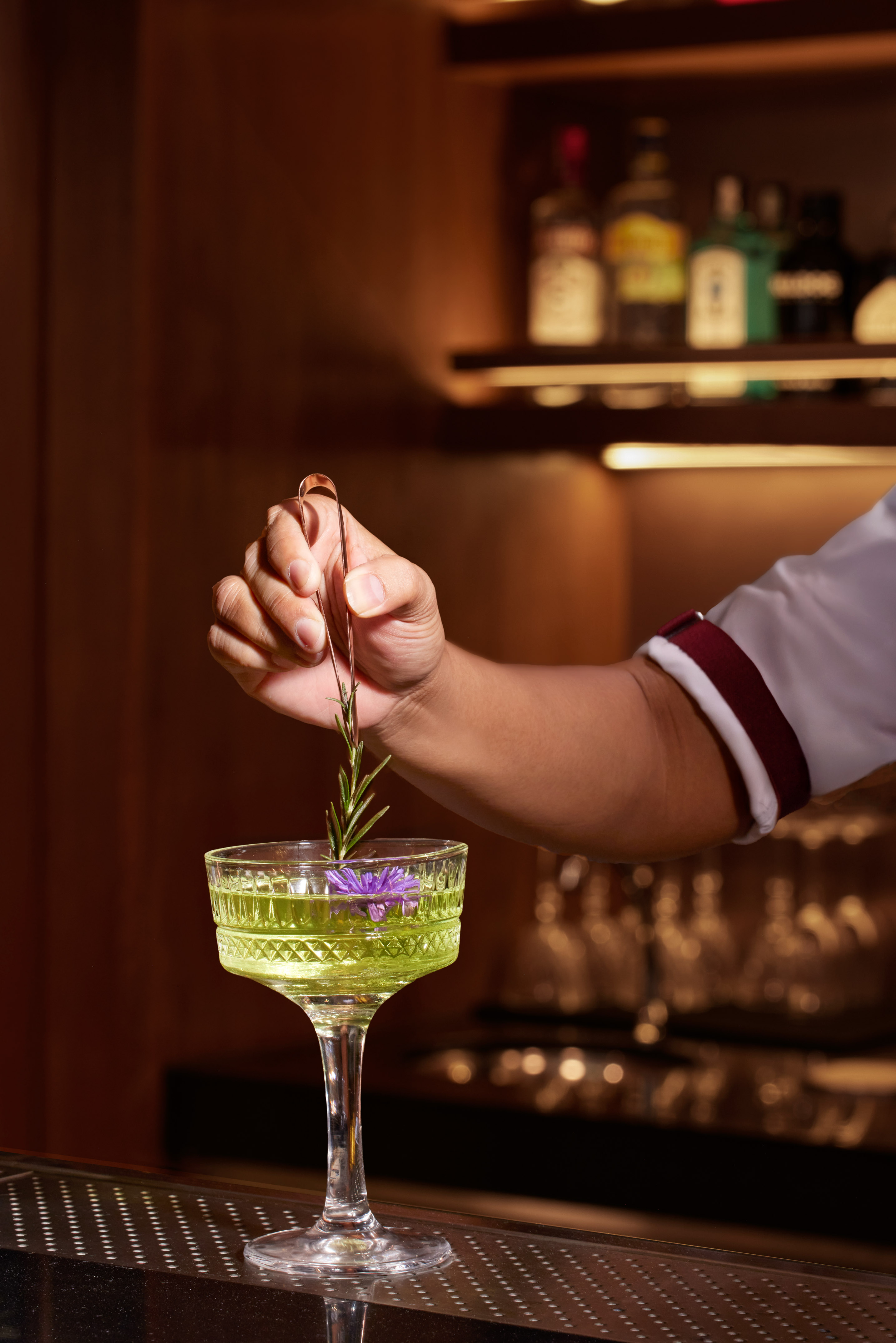 Bartender garnishing a vibrant green cocktail with rosemary and a purple flower.