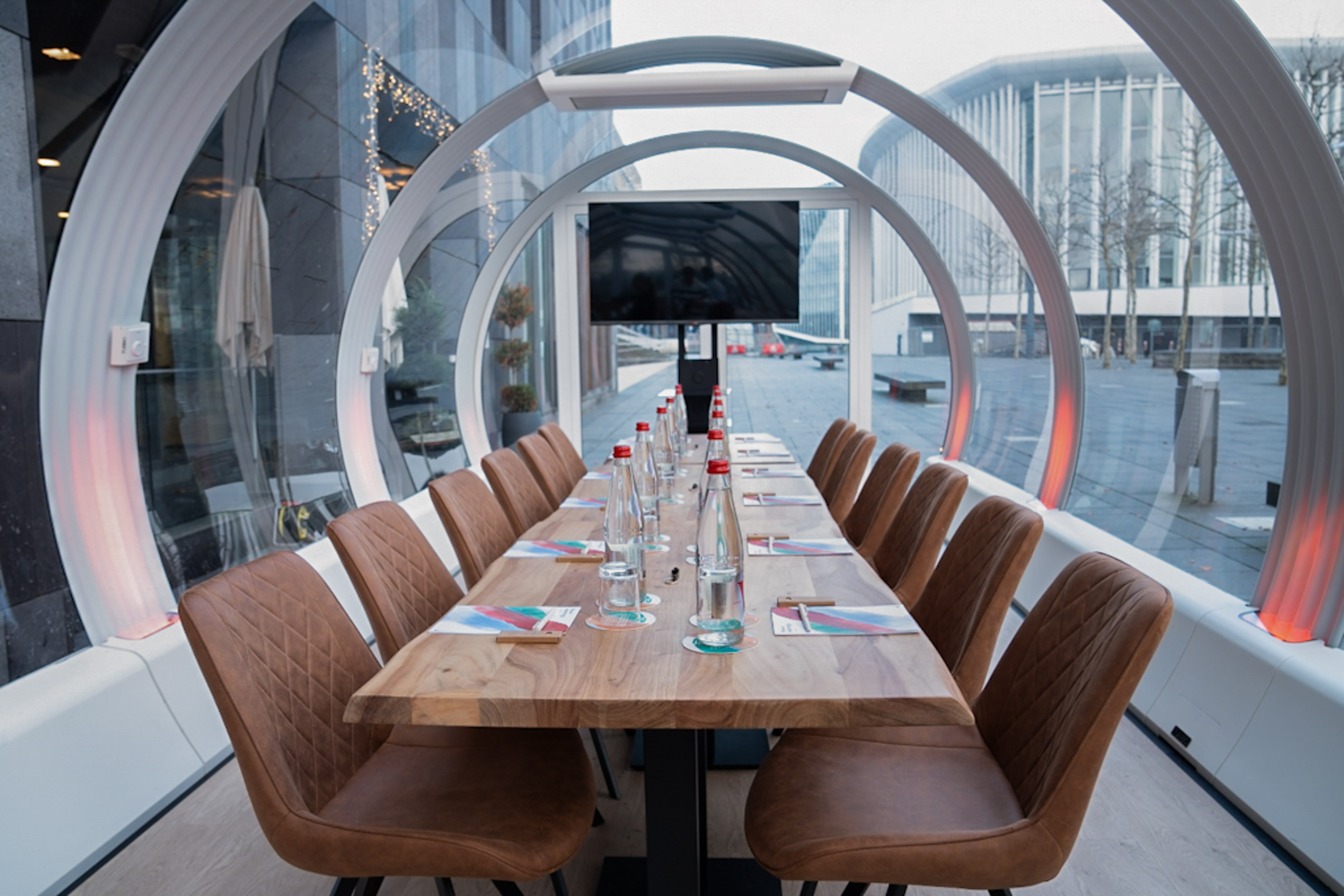 Glass conference room with wood table, brown chairs, and city view.