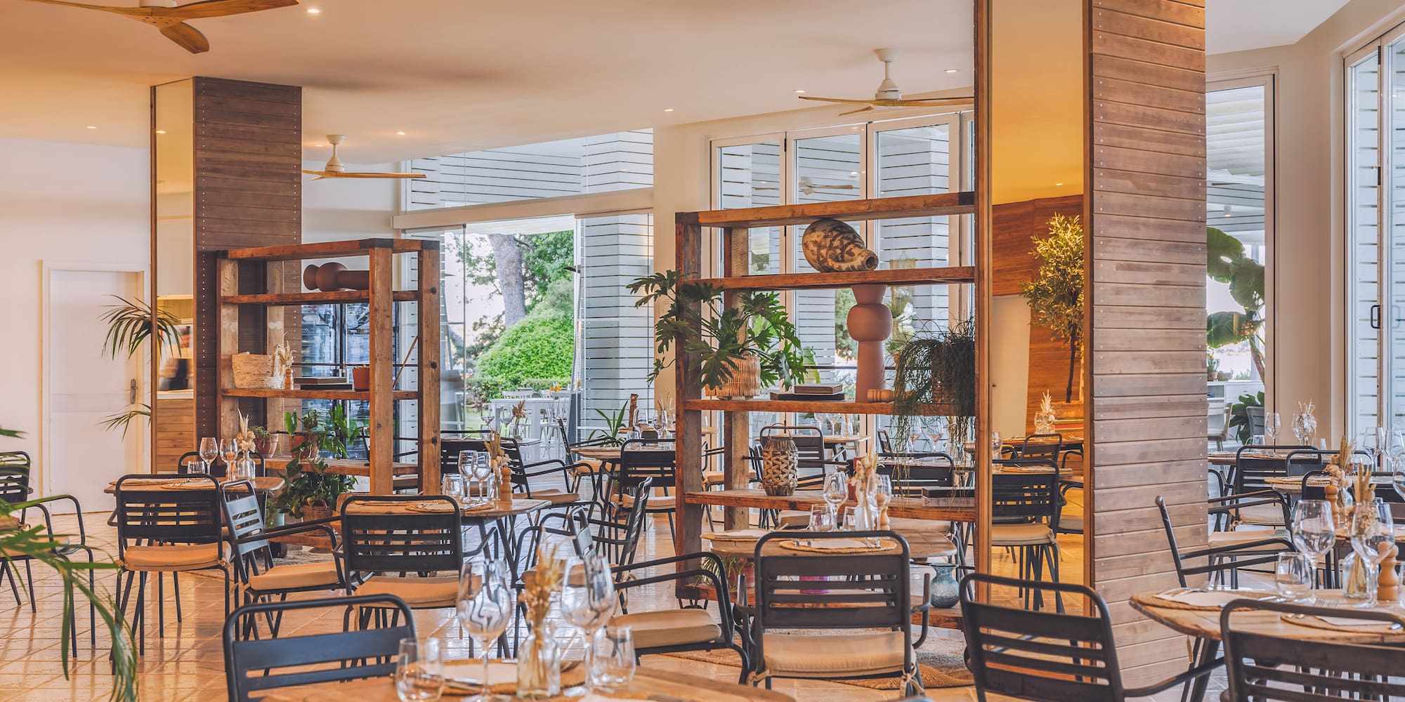 Modern restaurant dining area with wooden tables, dark chairs, and indoor plants.