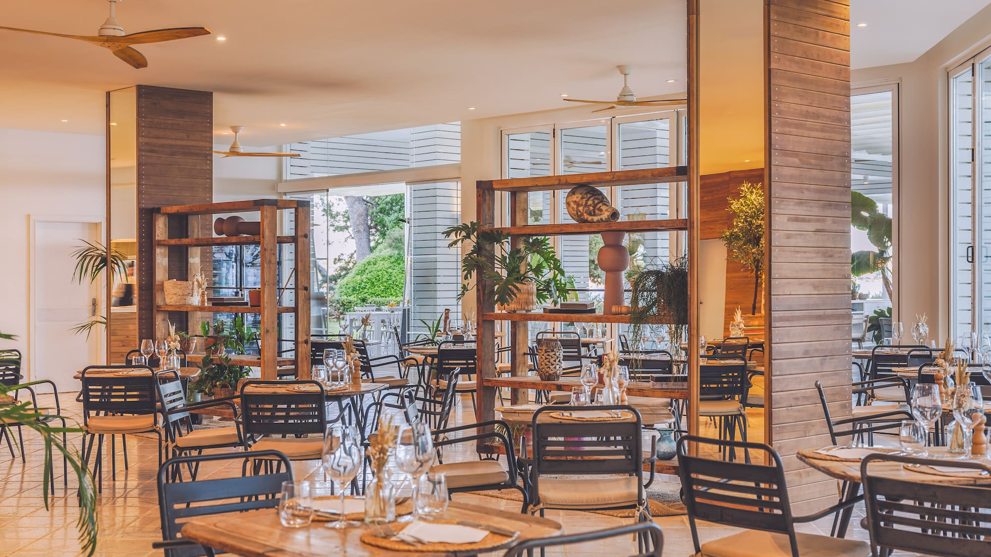 Modern restaurant dining area with wooden tables, dark chairs, and indoor plants.