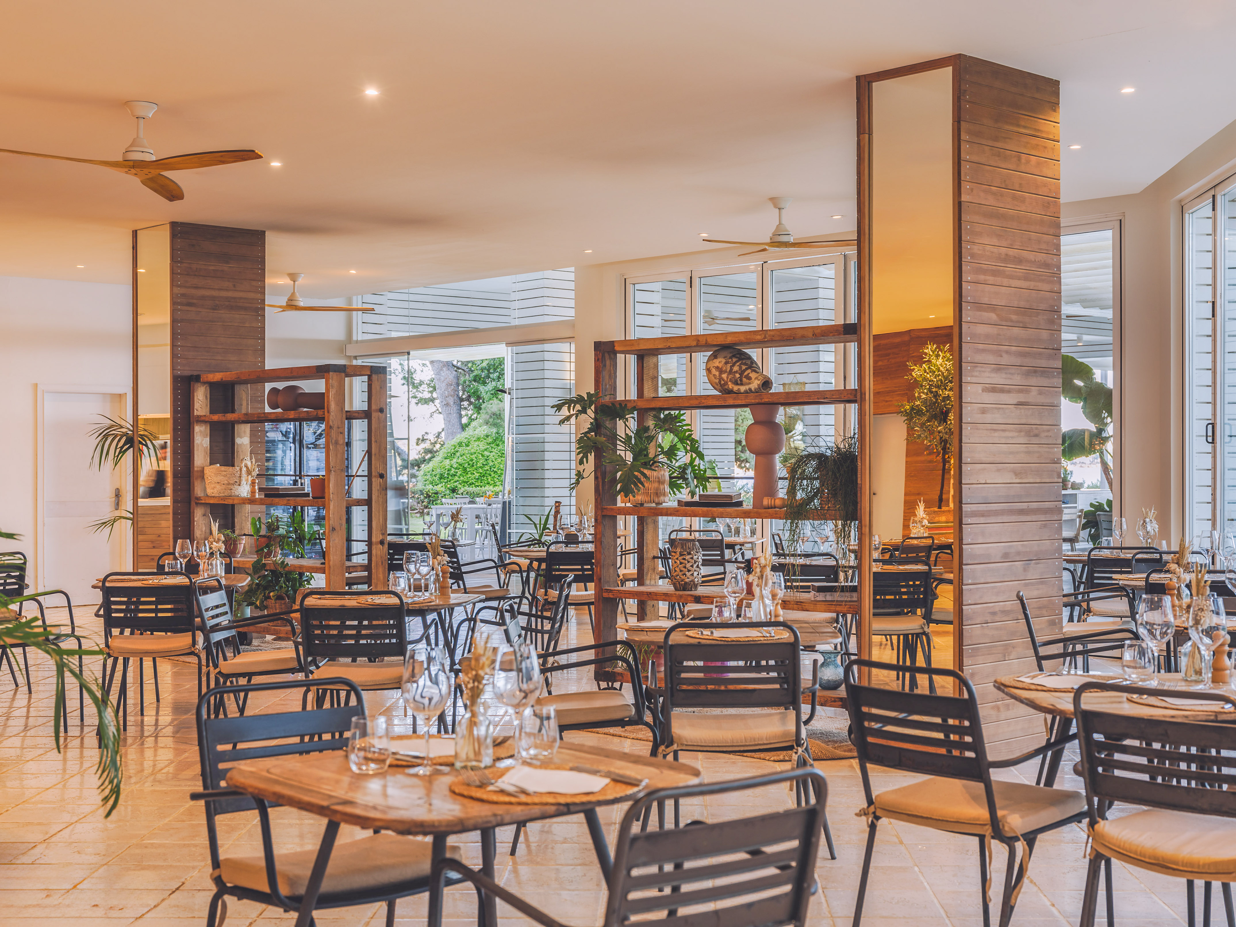 Modern restaurant dining area with wooden tables, dark chairs, and indoor plants.