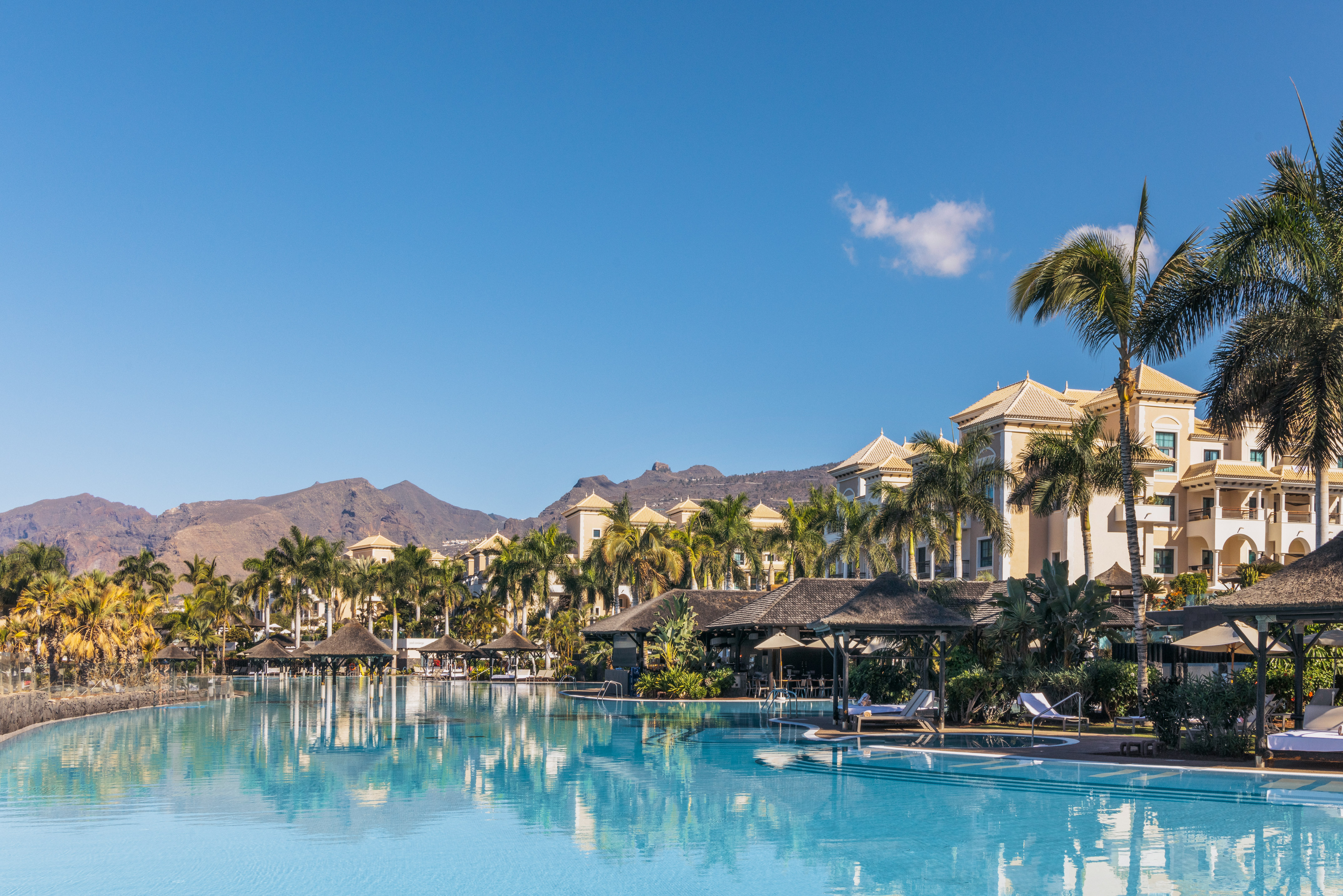 Tropical resort pool, palm trees, elegant hotel, and mountains.