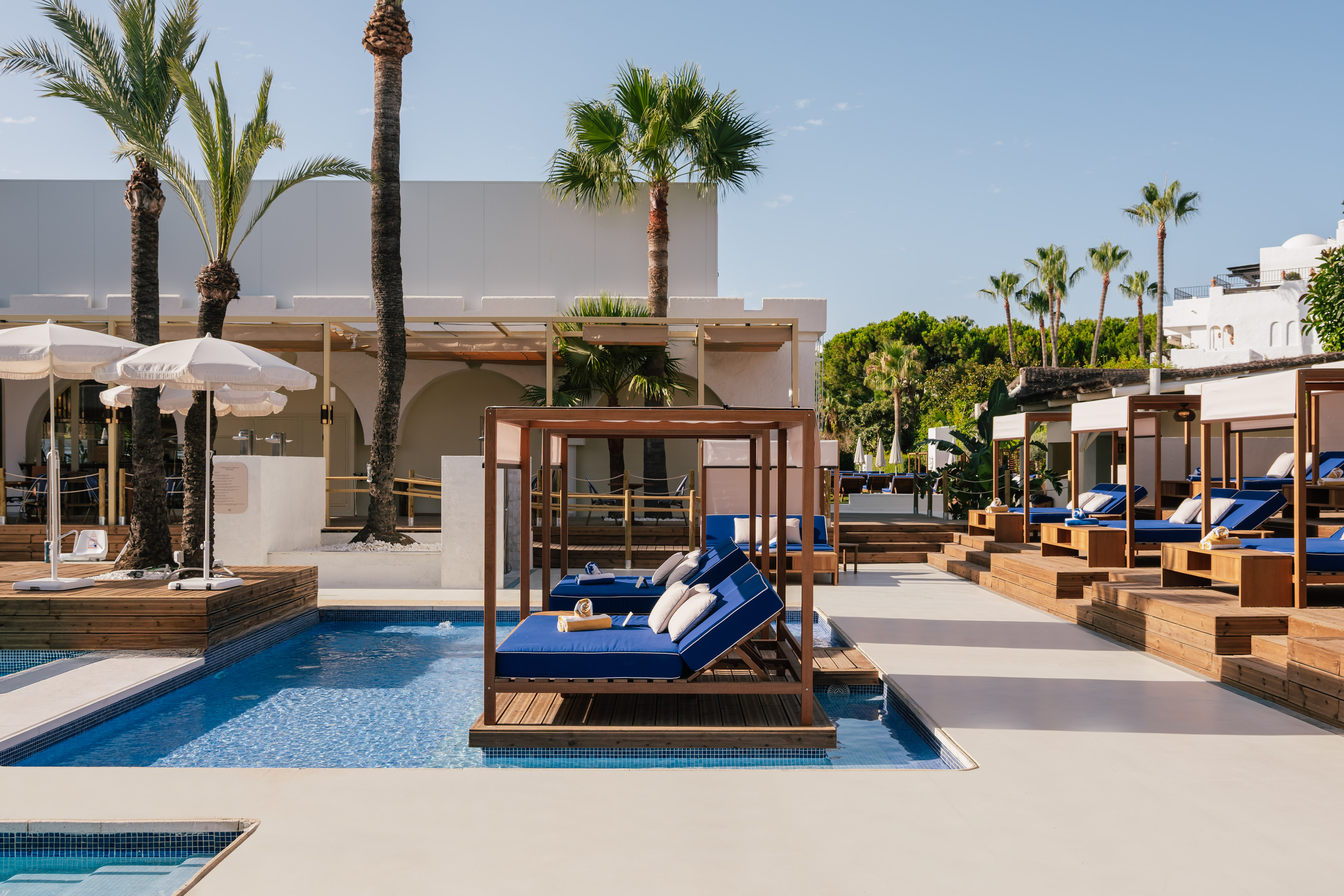 Luxury resort pool with wooden cabanas, blue daybeds, and palm trees.