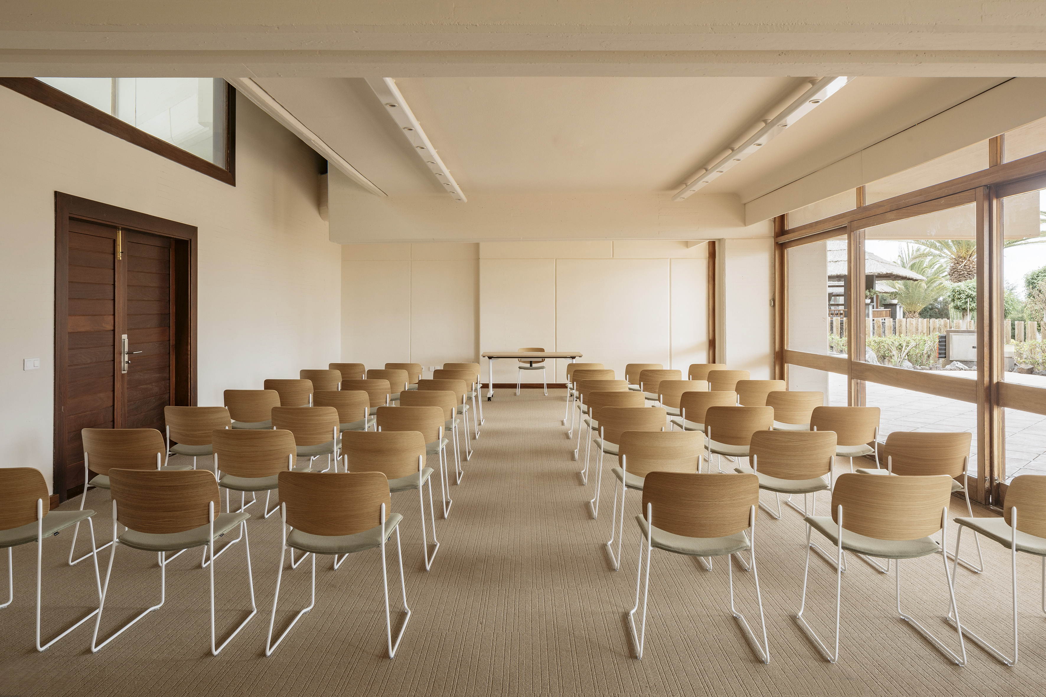 Modern meeting room, rows of wood chairs, front table, natural light, garden view.