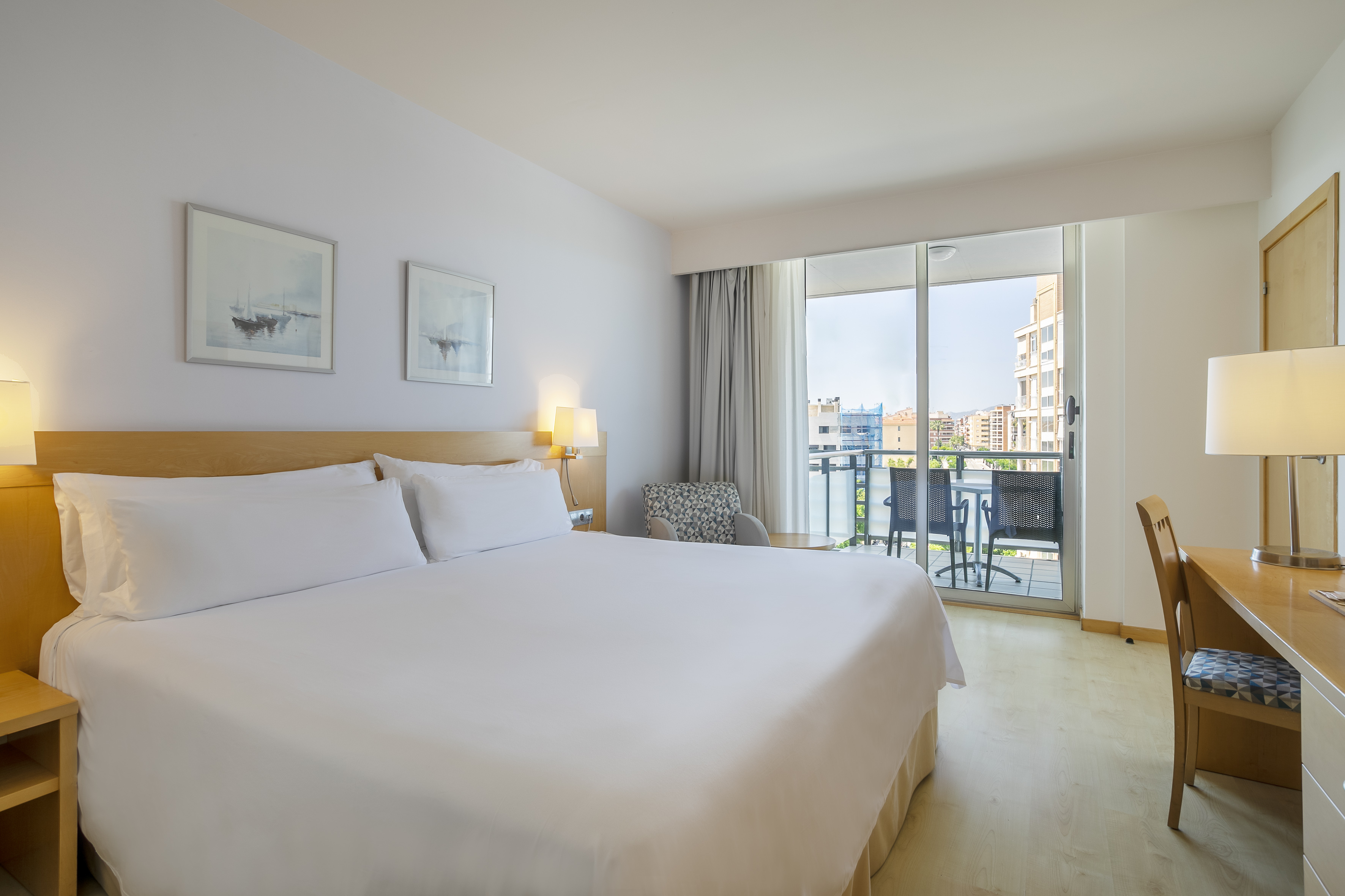 Modern hotel room with king bed, desk, and balcony cityscape view.
