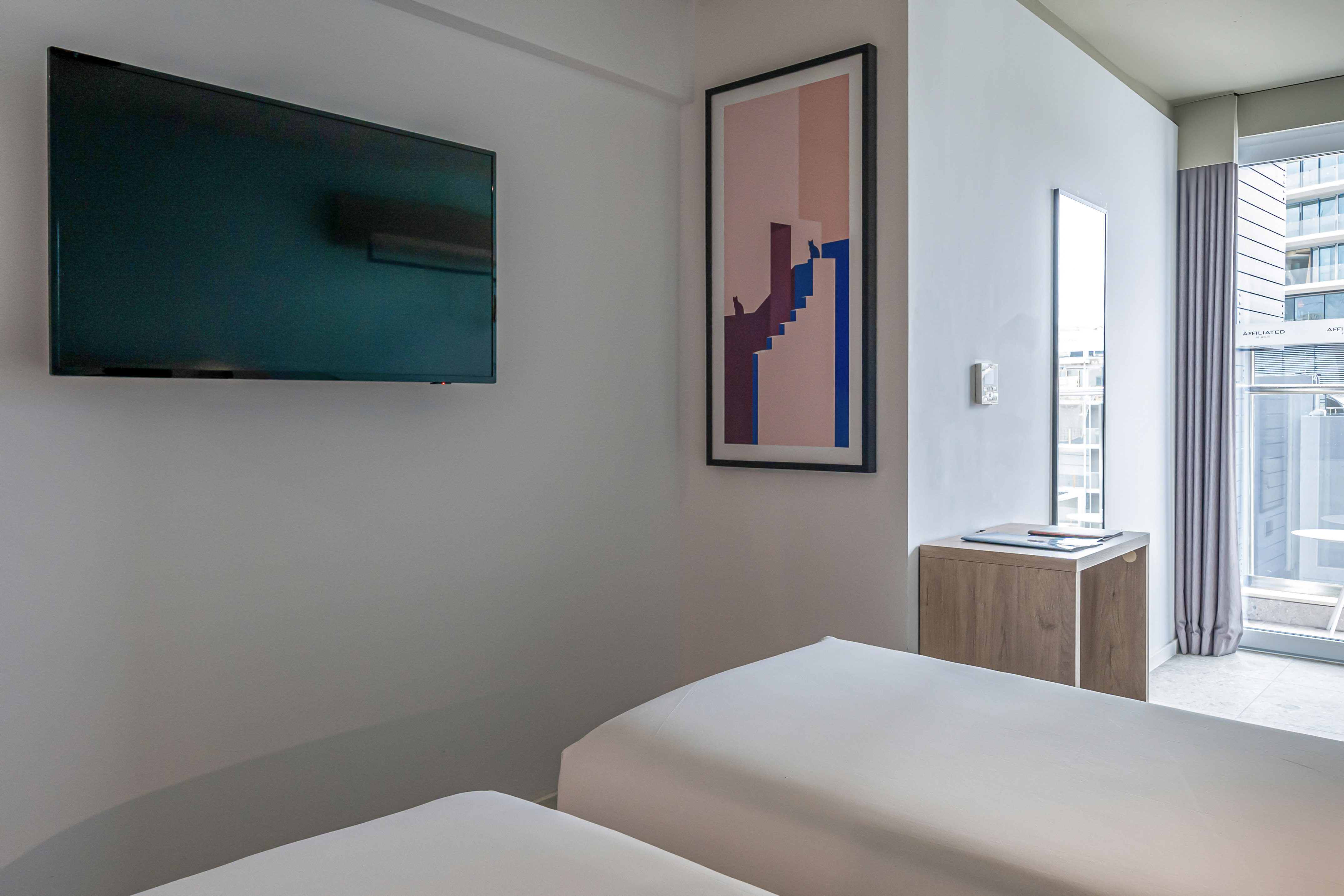 Modern hotel room with twin beds, wall-mounted TV, abstract art, and city view balcony.