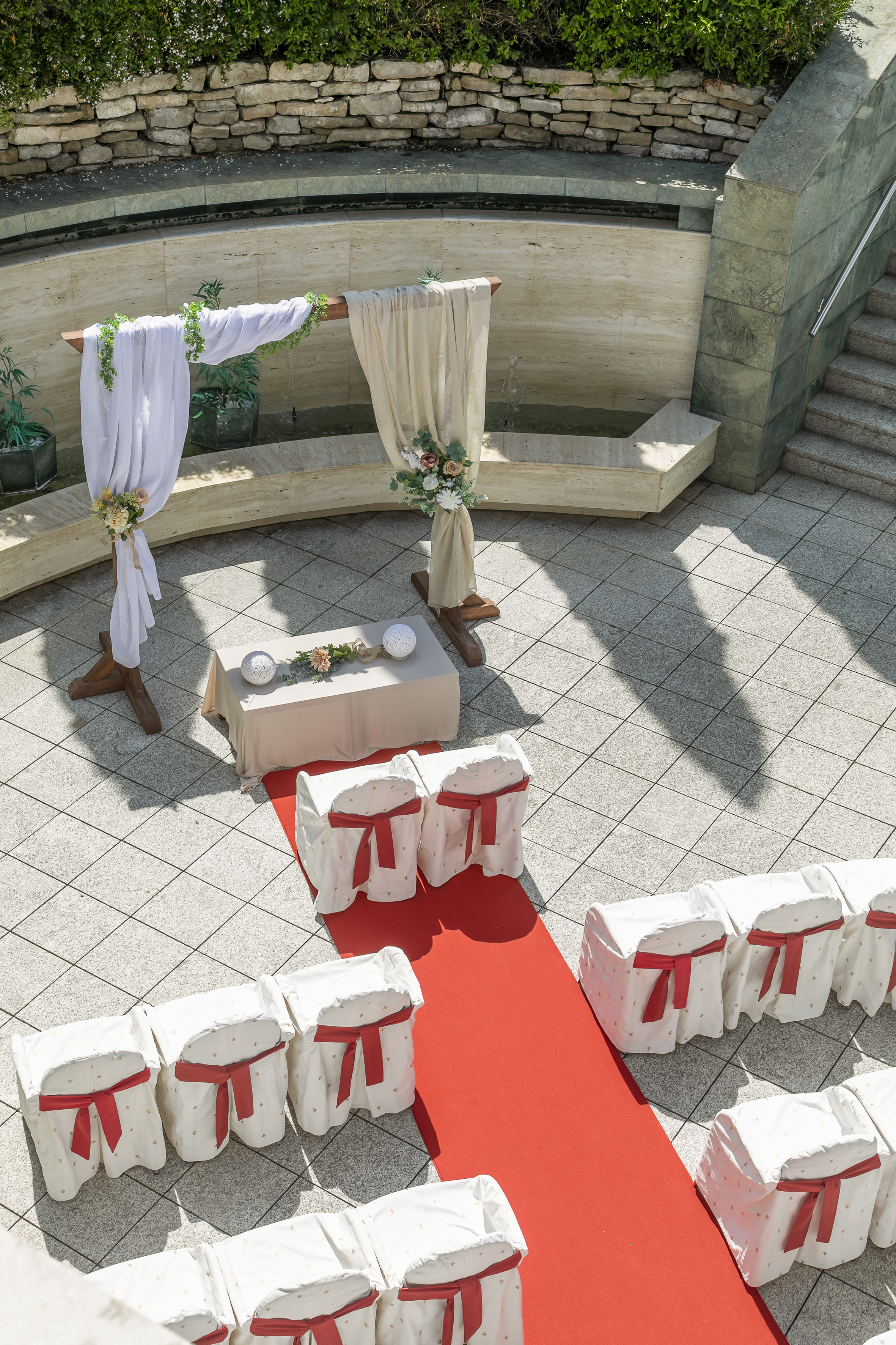 Outdoor wedding setup: floral arch, red carpet aisle, white chairs with red sashes.