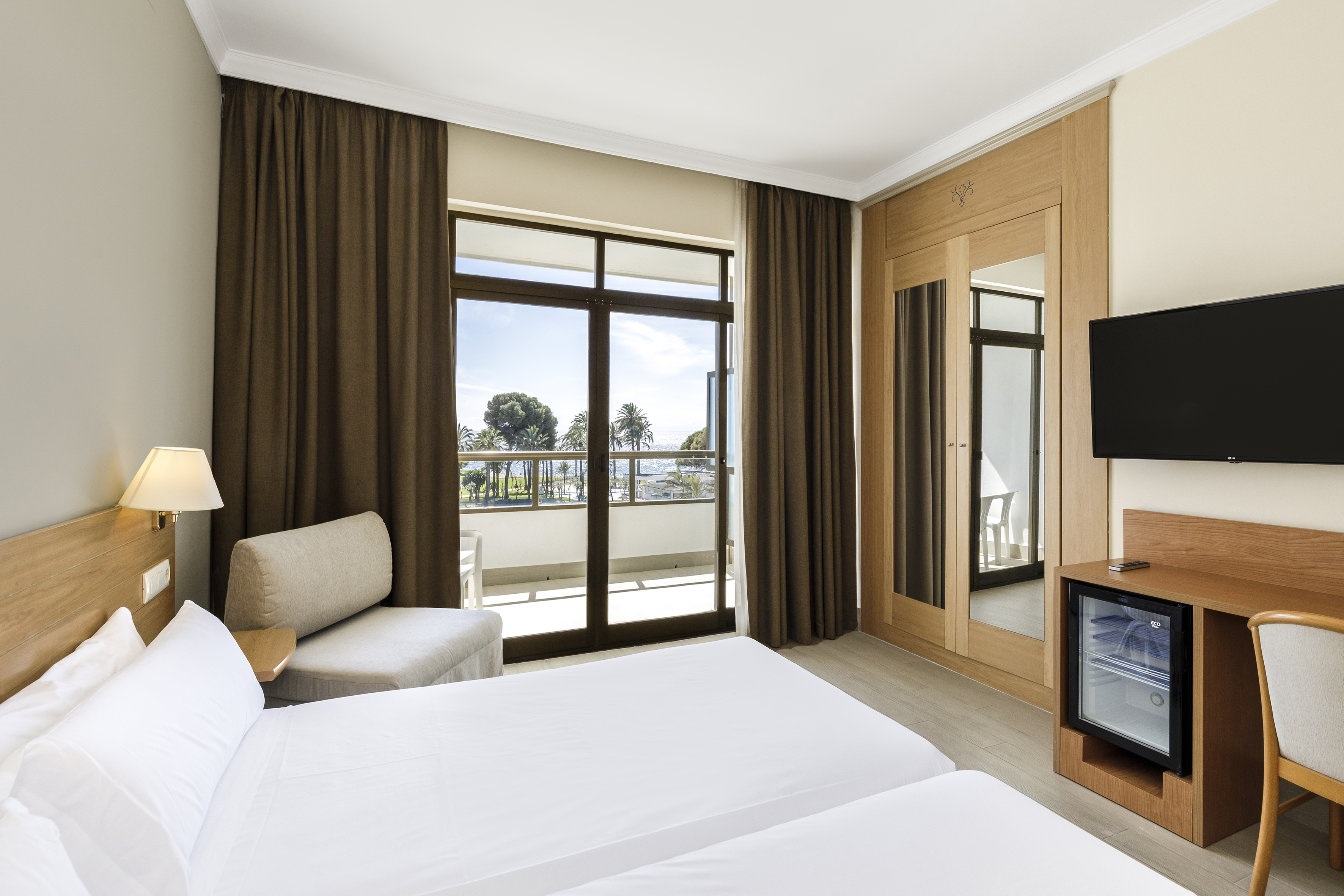 Hotel room with twin beds and balcony overlooking the sea with palm trees.