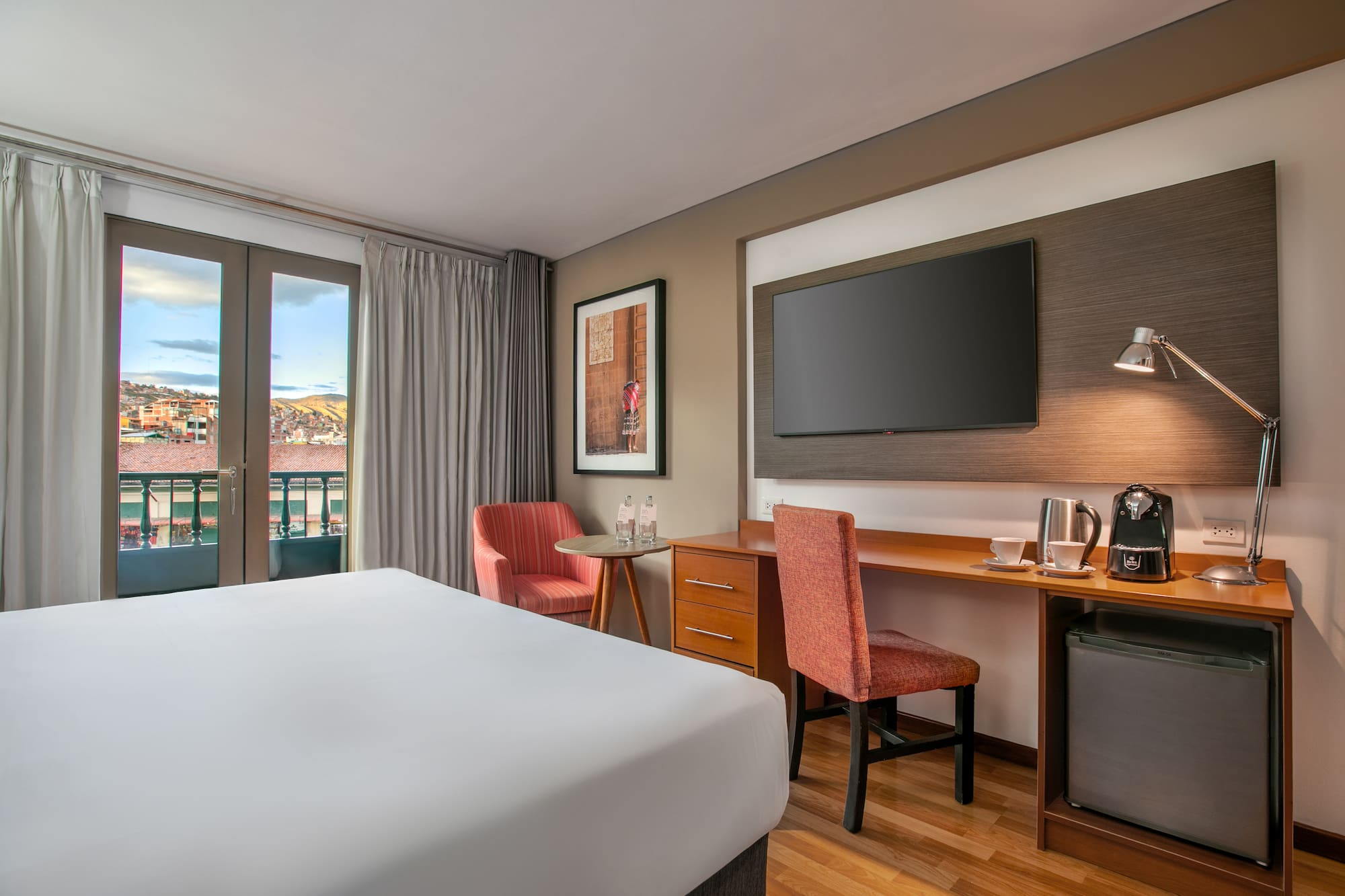 Cozy hotel room with a white bed, desk, TV, and balcony overlooking city rooftops. - Hotel Cusco La Paccha Affiliated by Meliá, Cusco