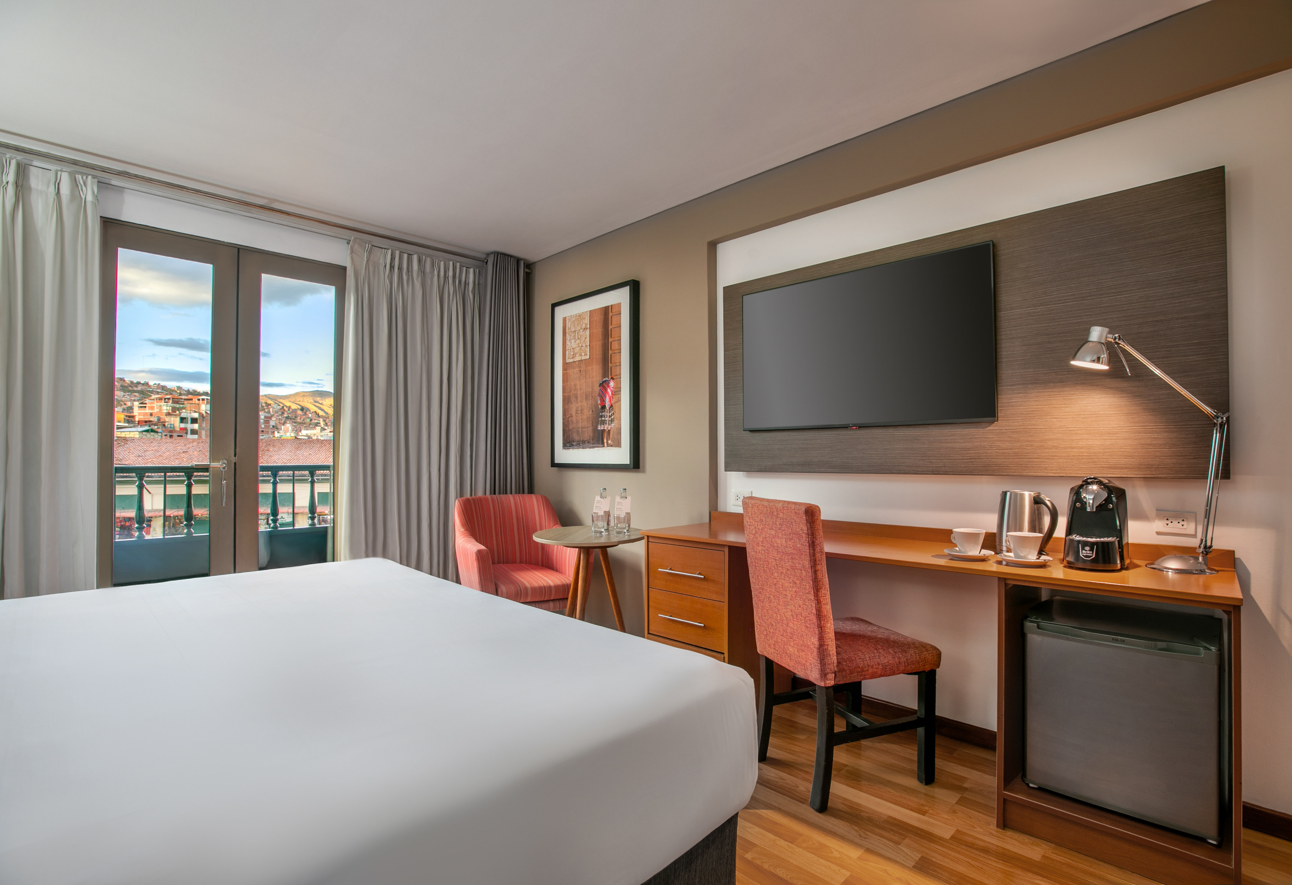 Cozy hotel room with a white bed, desk, TV, and balcony overlooking city rooftops. - Hotel Cusco La Paccha Affiliated by Meliá, Cusco