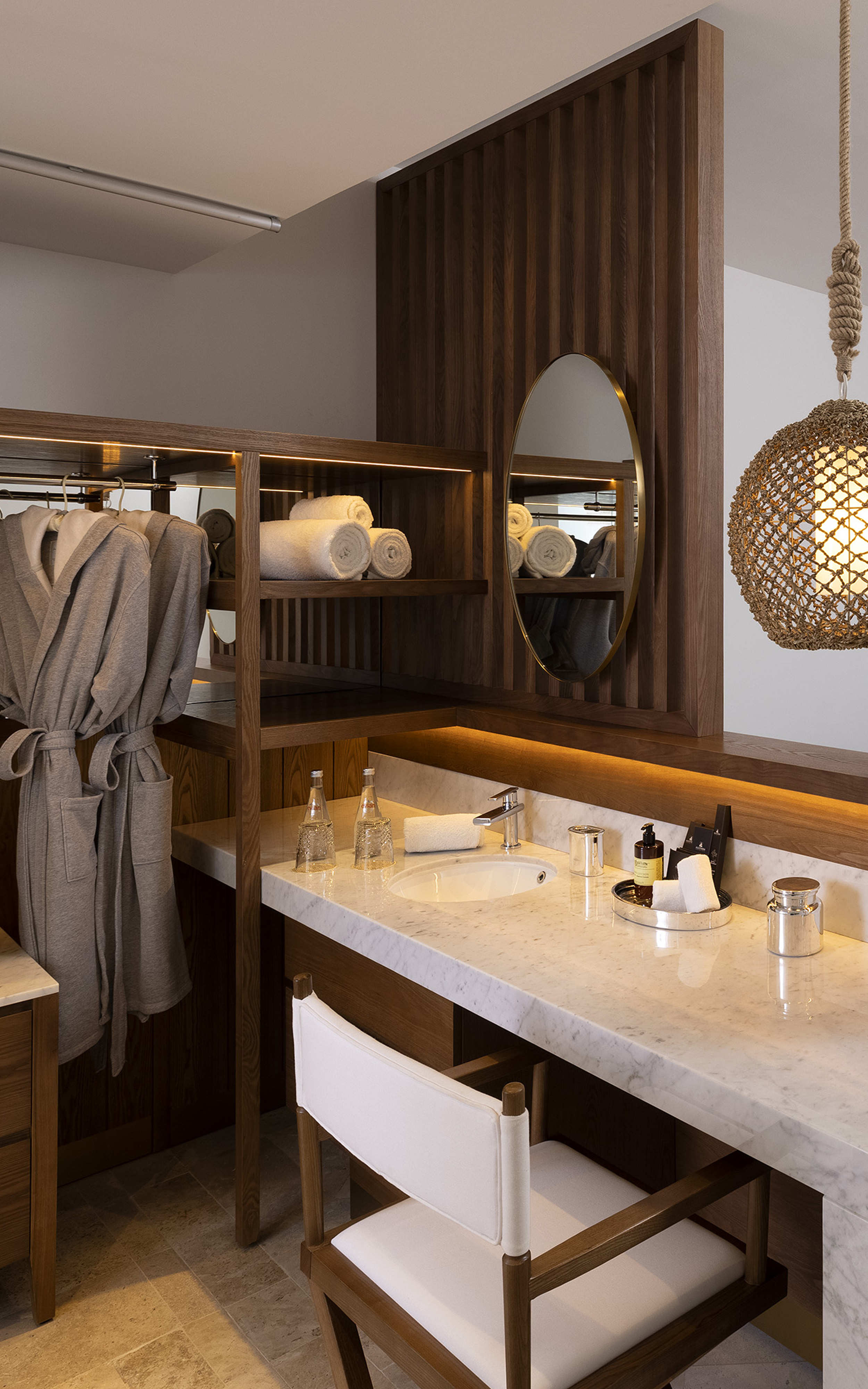 Hotel vanity area: marble counter, wood shelving, towels, bathrobe, oval mirror.