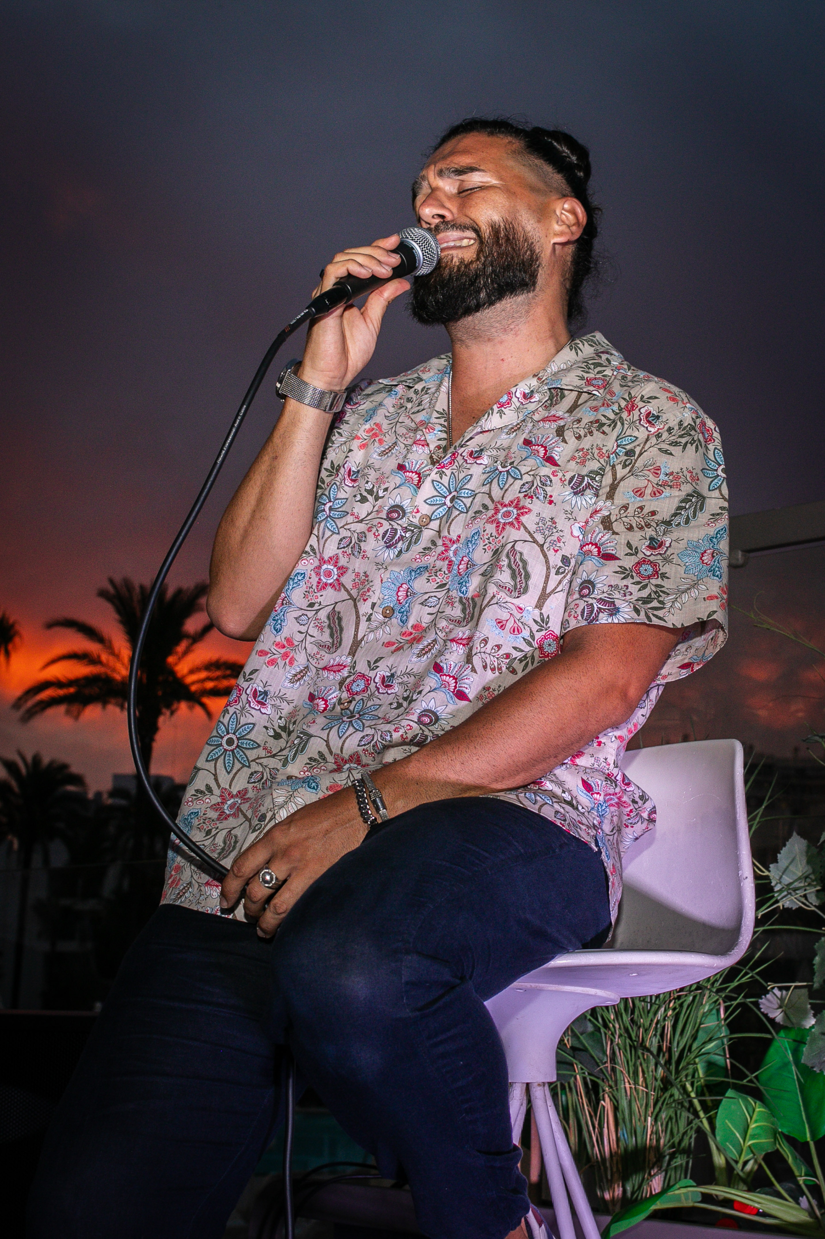 Bearded singer in floral shirt performs with microphone during sunset.
