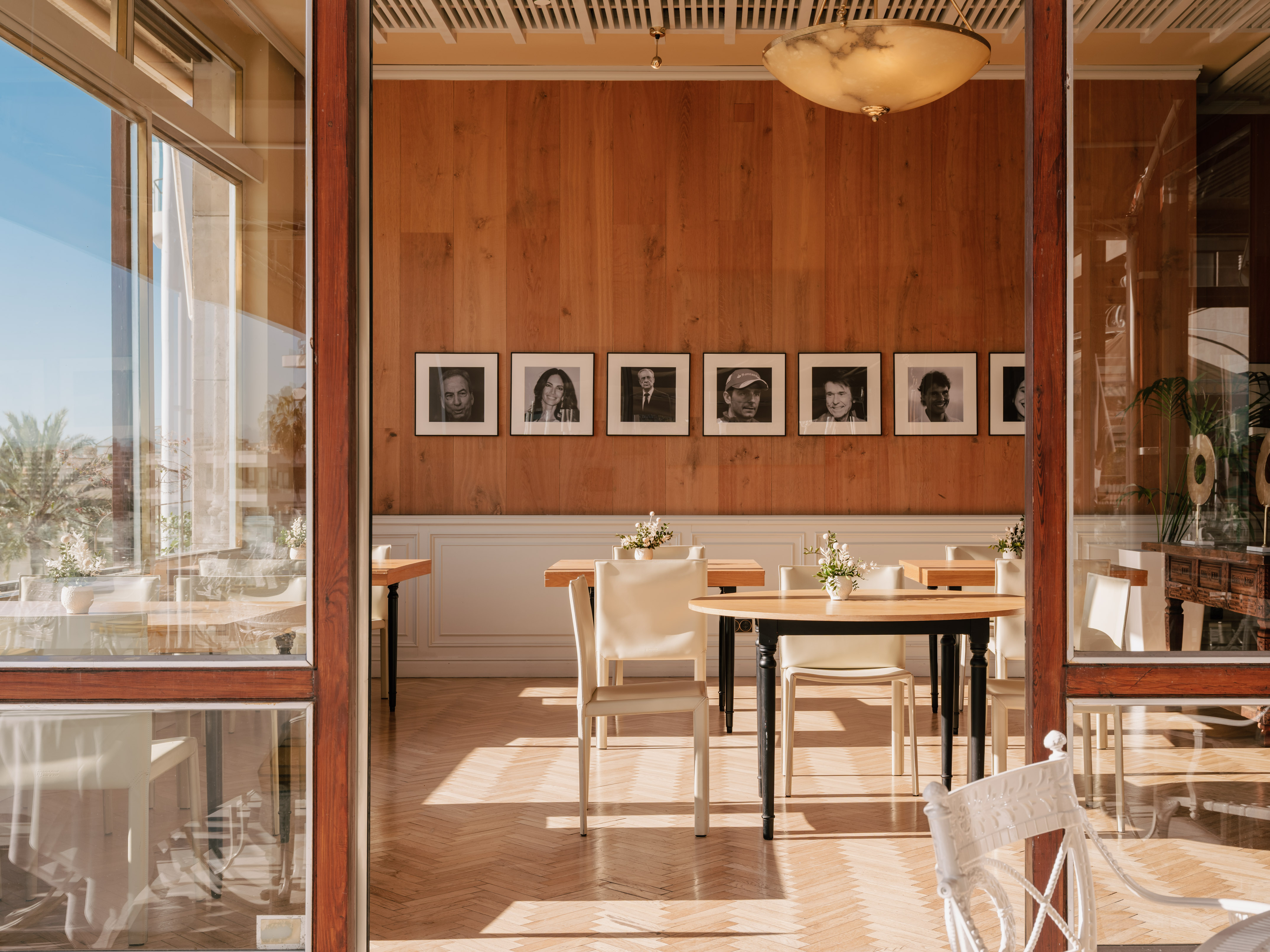 Sunlit restaurant, wooden walls with framed portraits, dining tables, and herringbone floor.