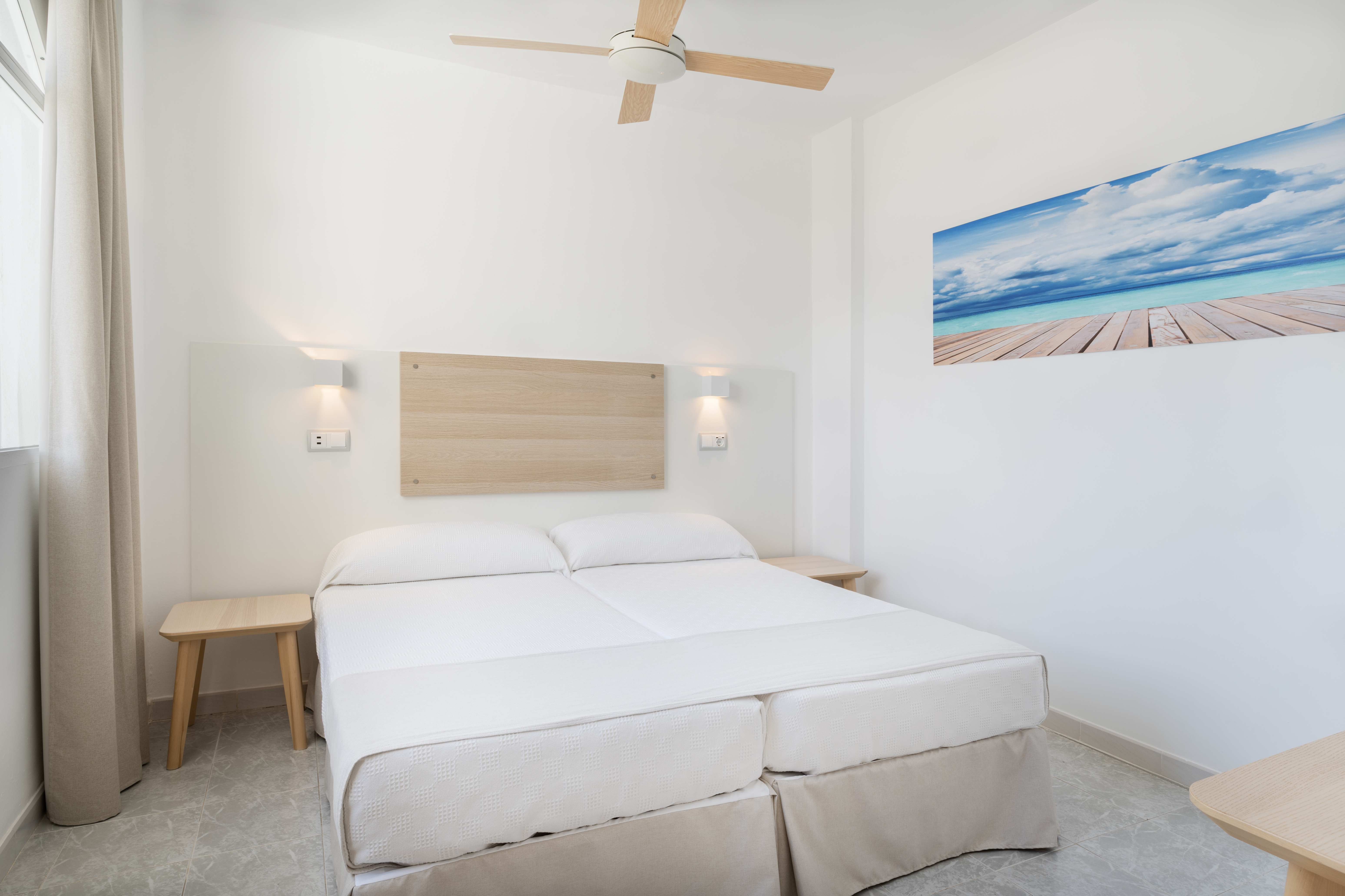 Modern hotel bedroom with white twin beds, wooden headboard, ceiling fan, ocean art.