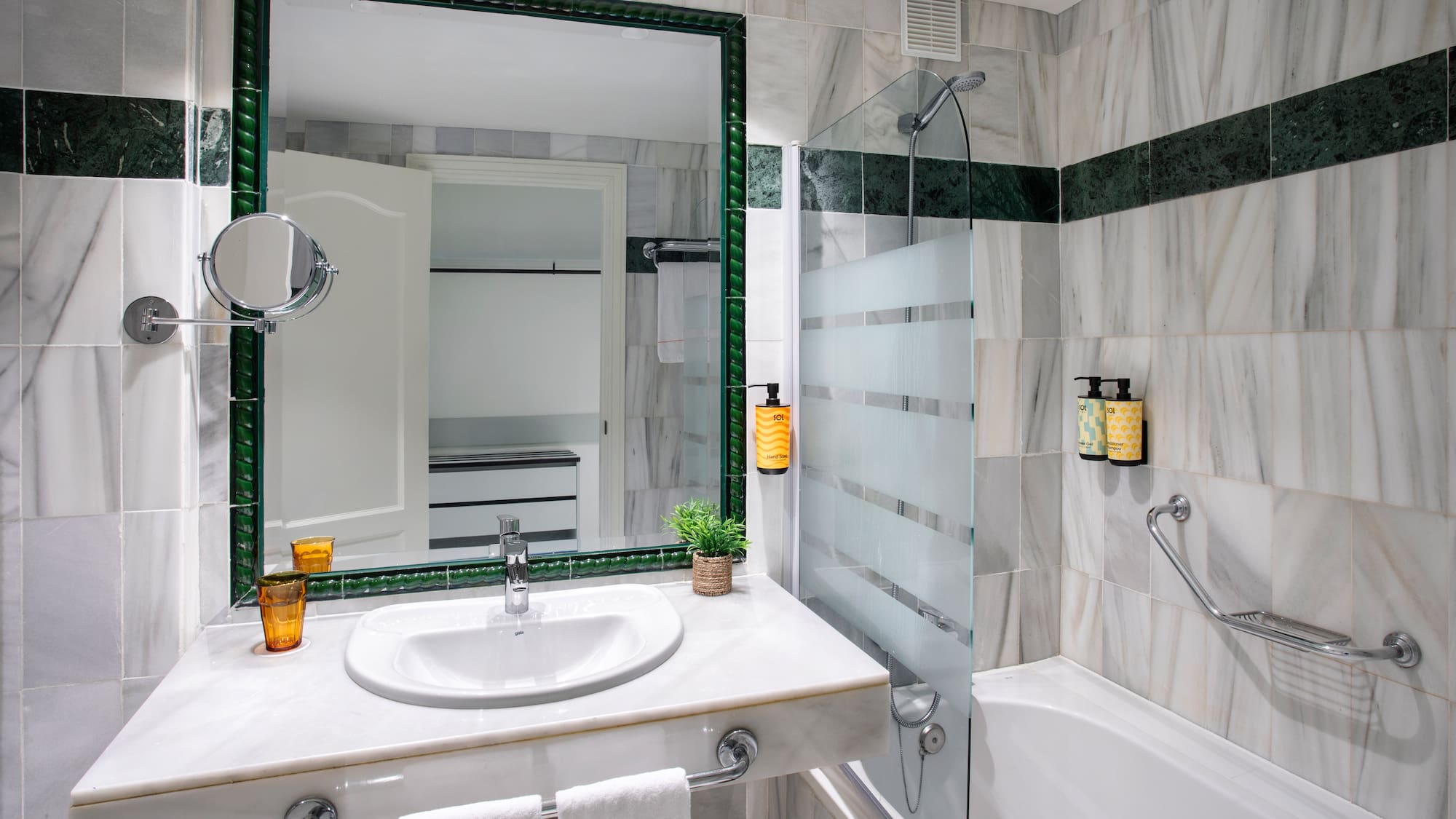 Baño elegante con paredes de mármol y detalles verdes, lavabo, espejo y ducha.