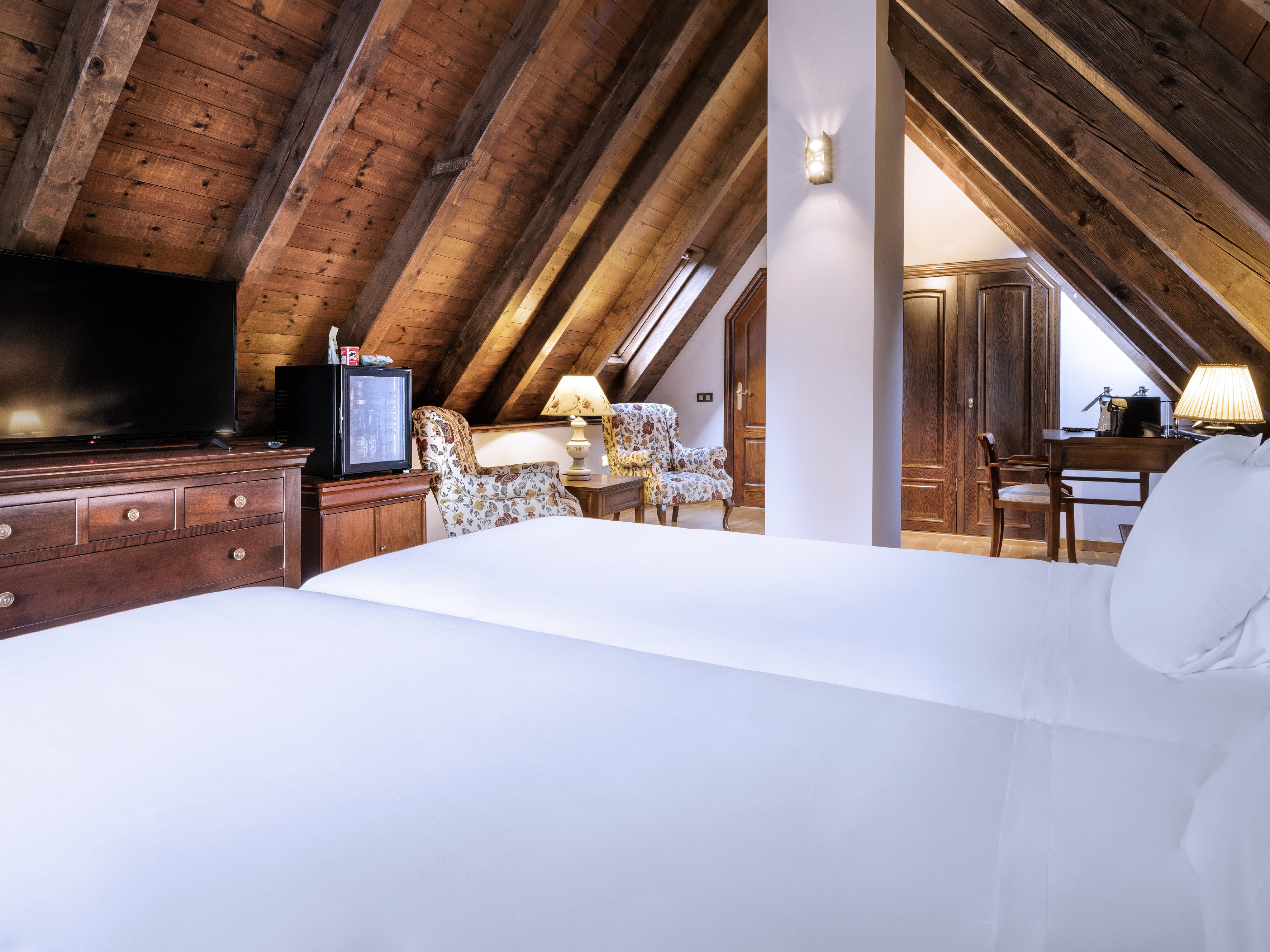 Attic hotel room with twin beds, beamed wooden ceiling, TV, and armchairs.