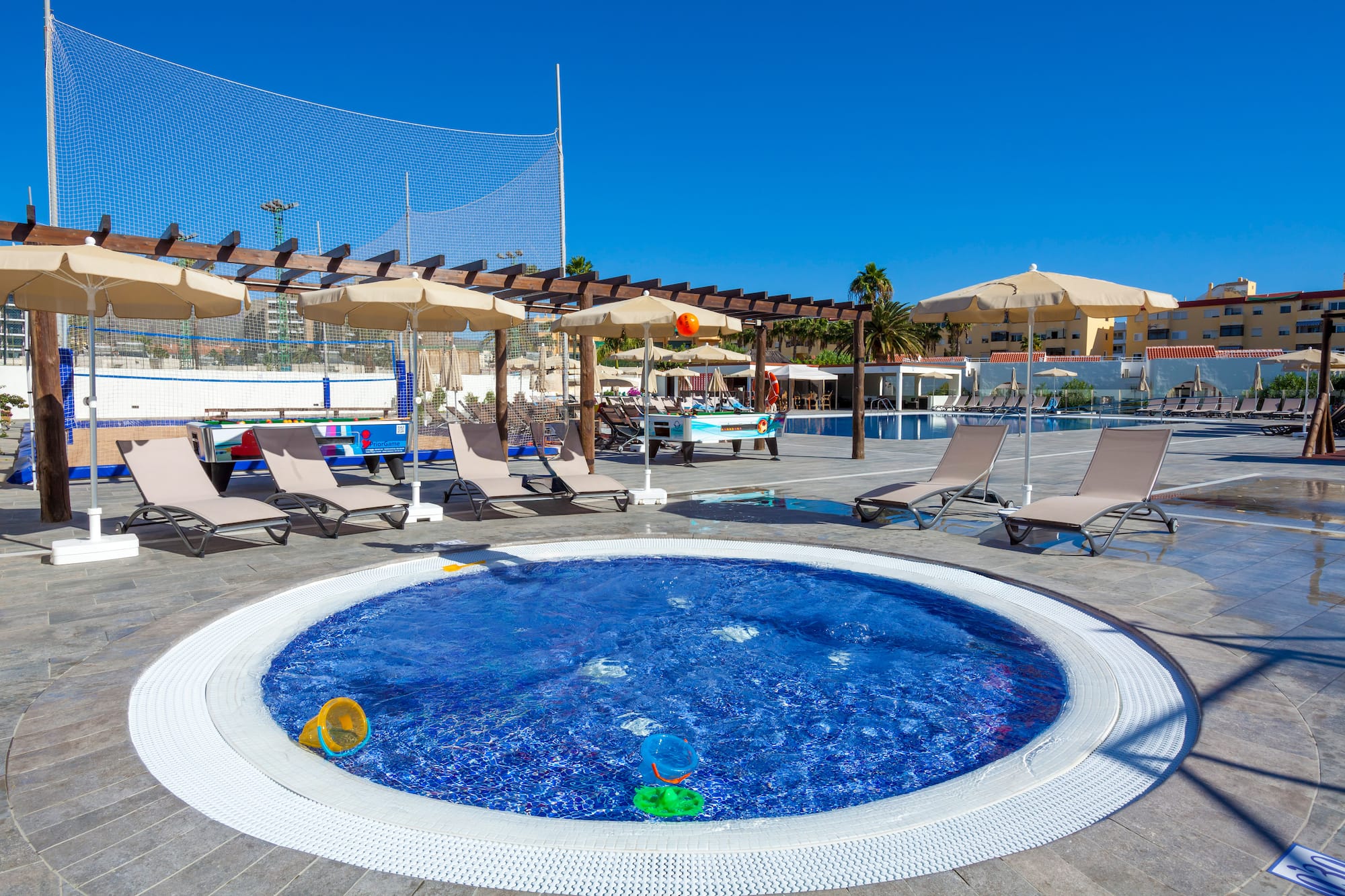 Sunny resort pool area with hot tub, lounge chairs, umbrellas, and outdoor games.