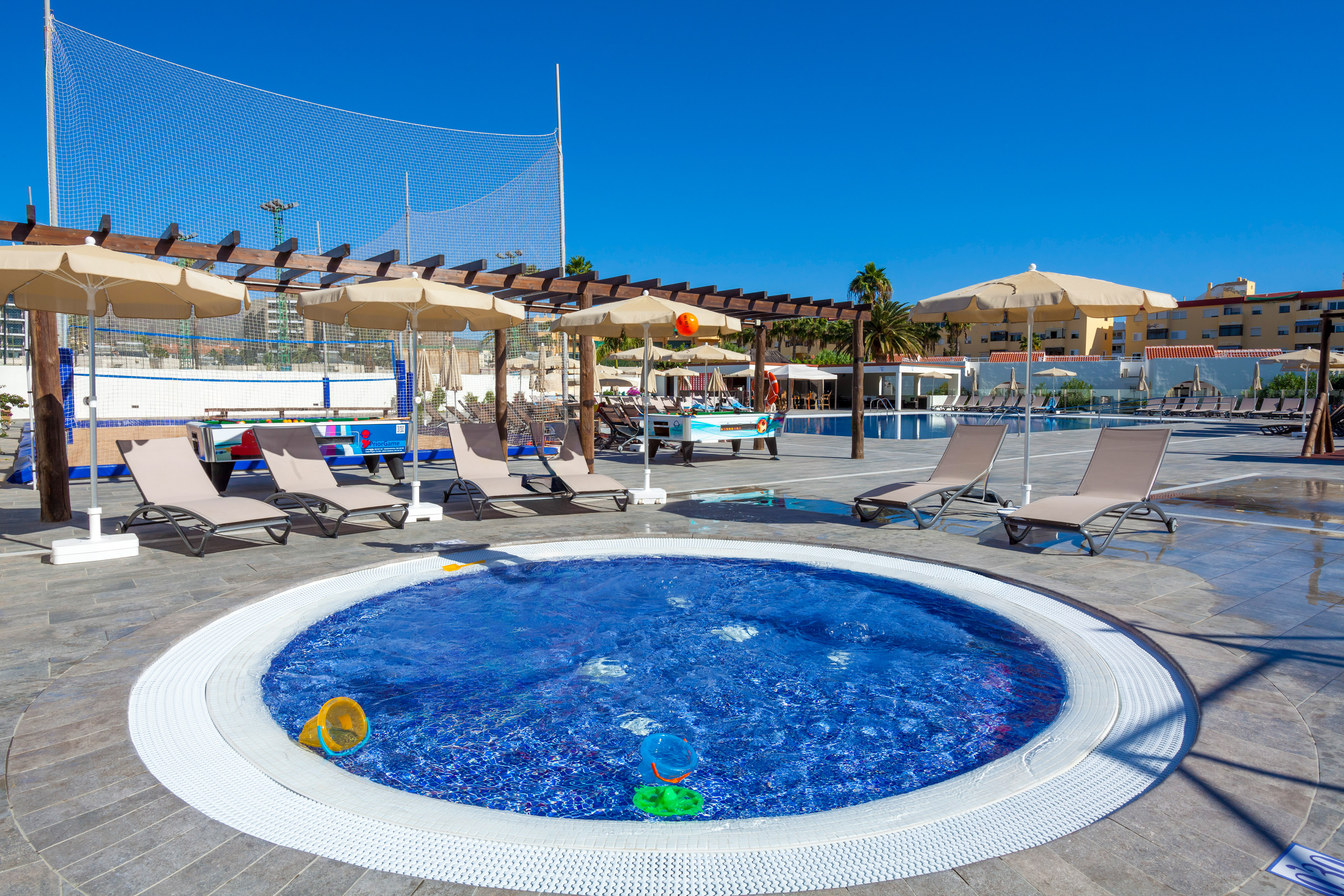 Sunny resort pool area with hot tub, lounge chairs, umbrellas, and outdoor games.