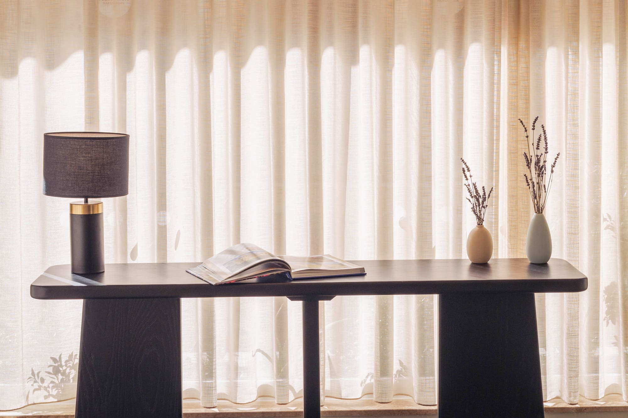 Black console table with lamp, open book, lavender vases. Sheer curtains, sunlit room.