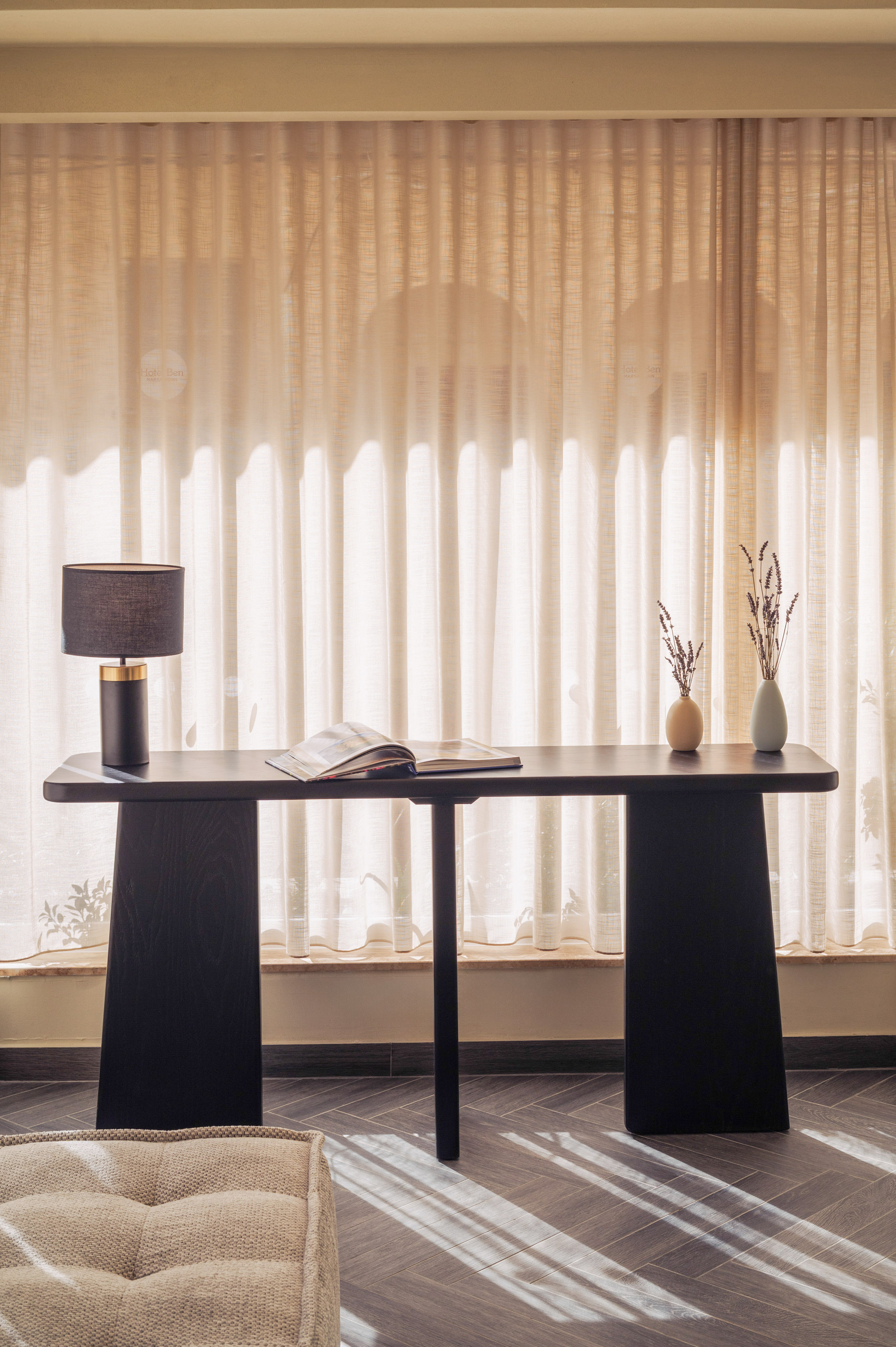 Black console table with lamp, open book, lavender vases. Sheer curtains, sunlit room.