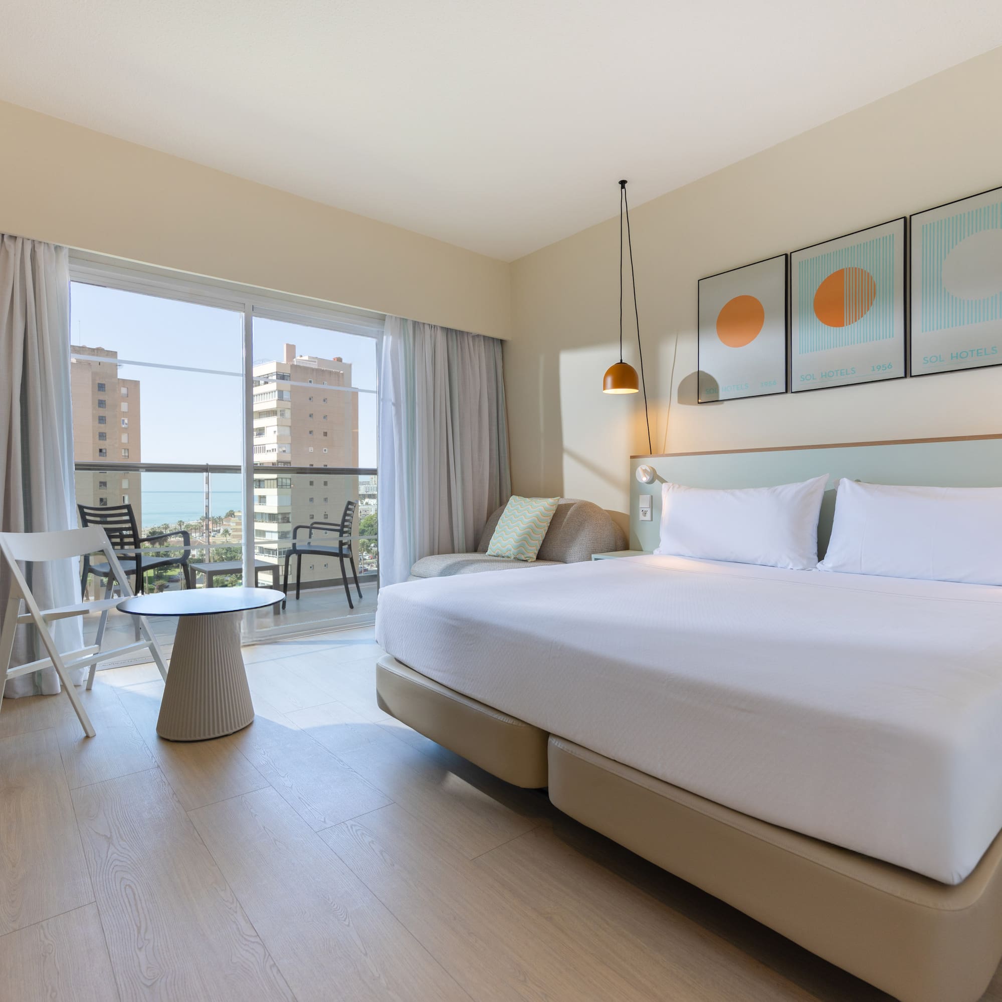 Modern hotel room with large bed and balcony overlooking sea and city.