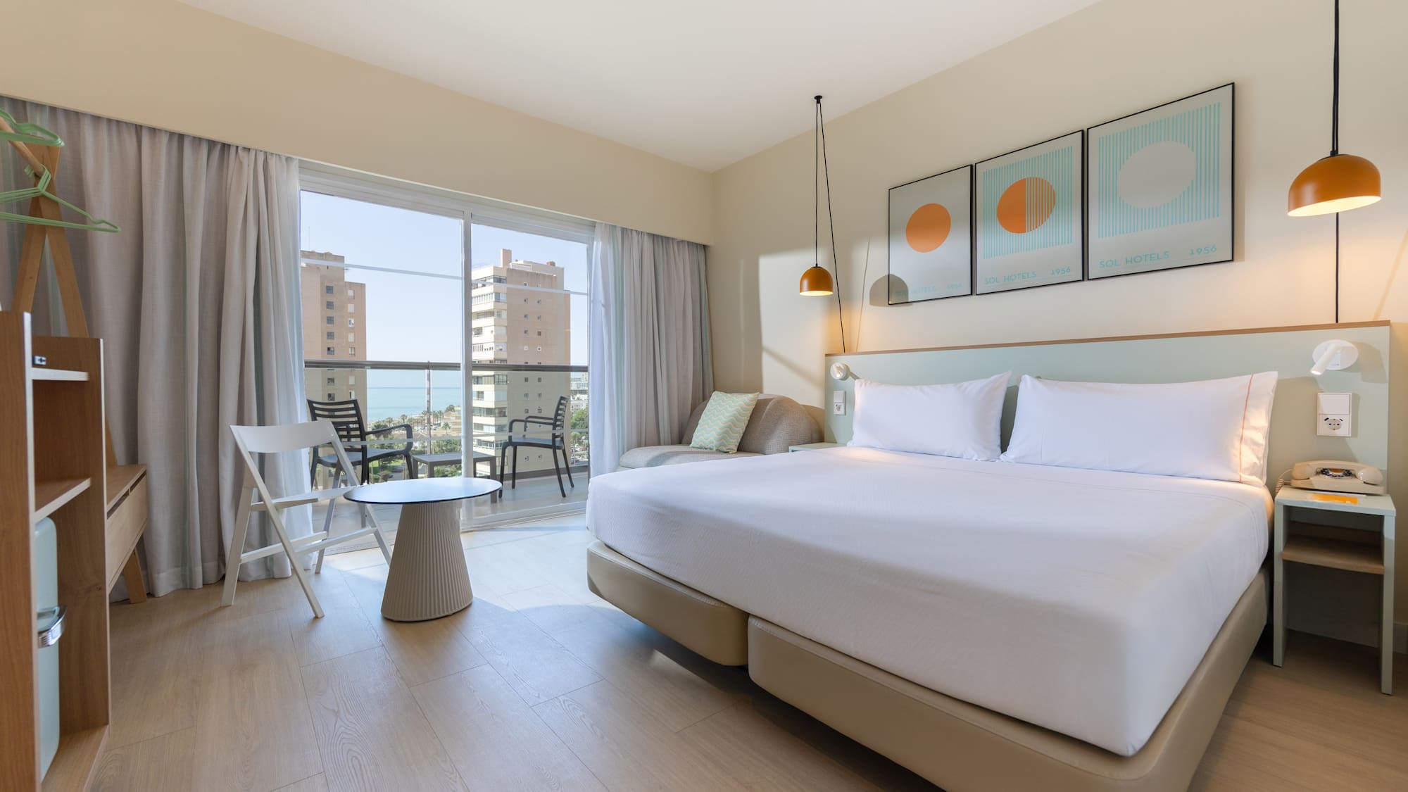Modern hotel room with large bed and balcony overlooking sea and city.