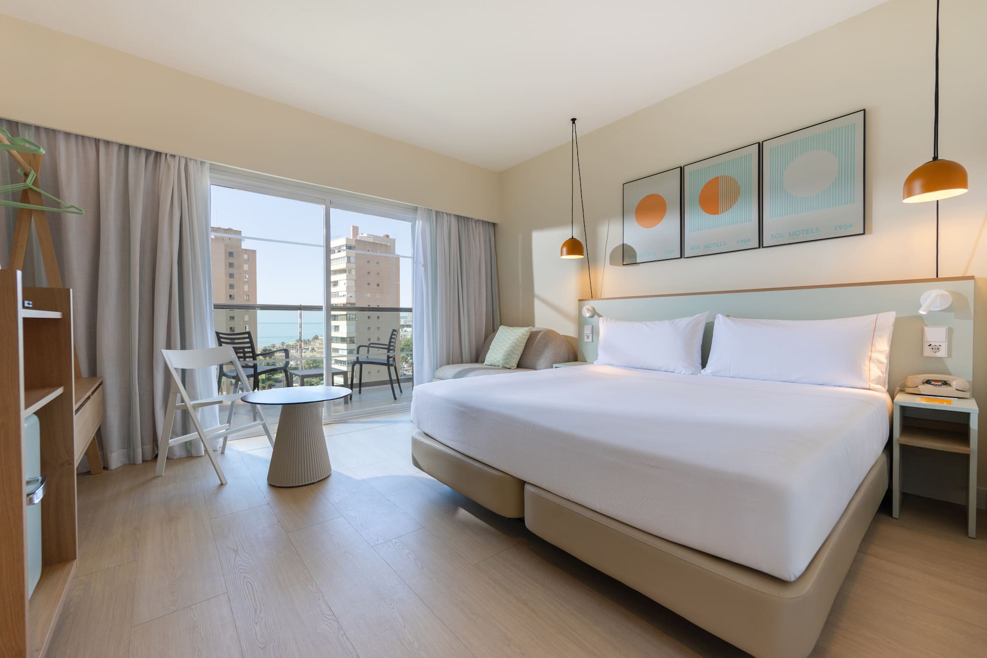 Modern hotel room with balcony ocean view, large bed, and light wood floor.