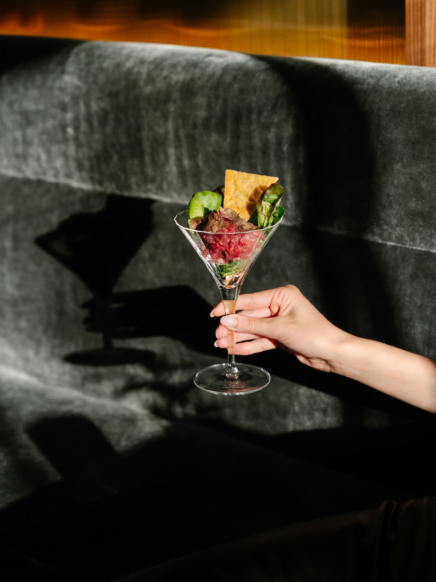 Martini glass with beef tartare, greens, and crispy chip, held by hand.