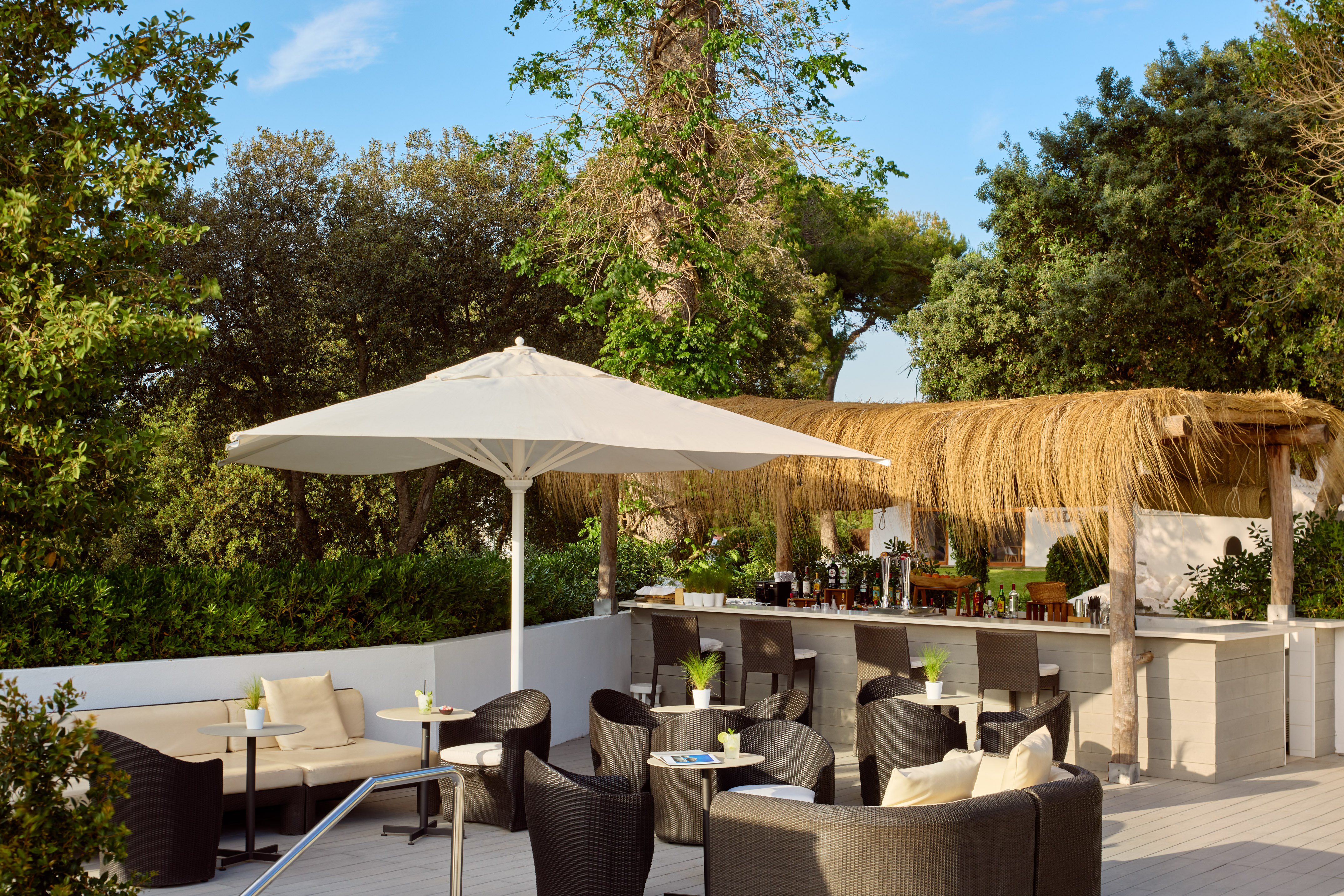 Outdoor bar with thatched roof, white umbrella, wicker seating, and green foliage.