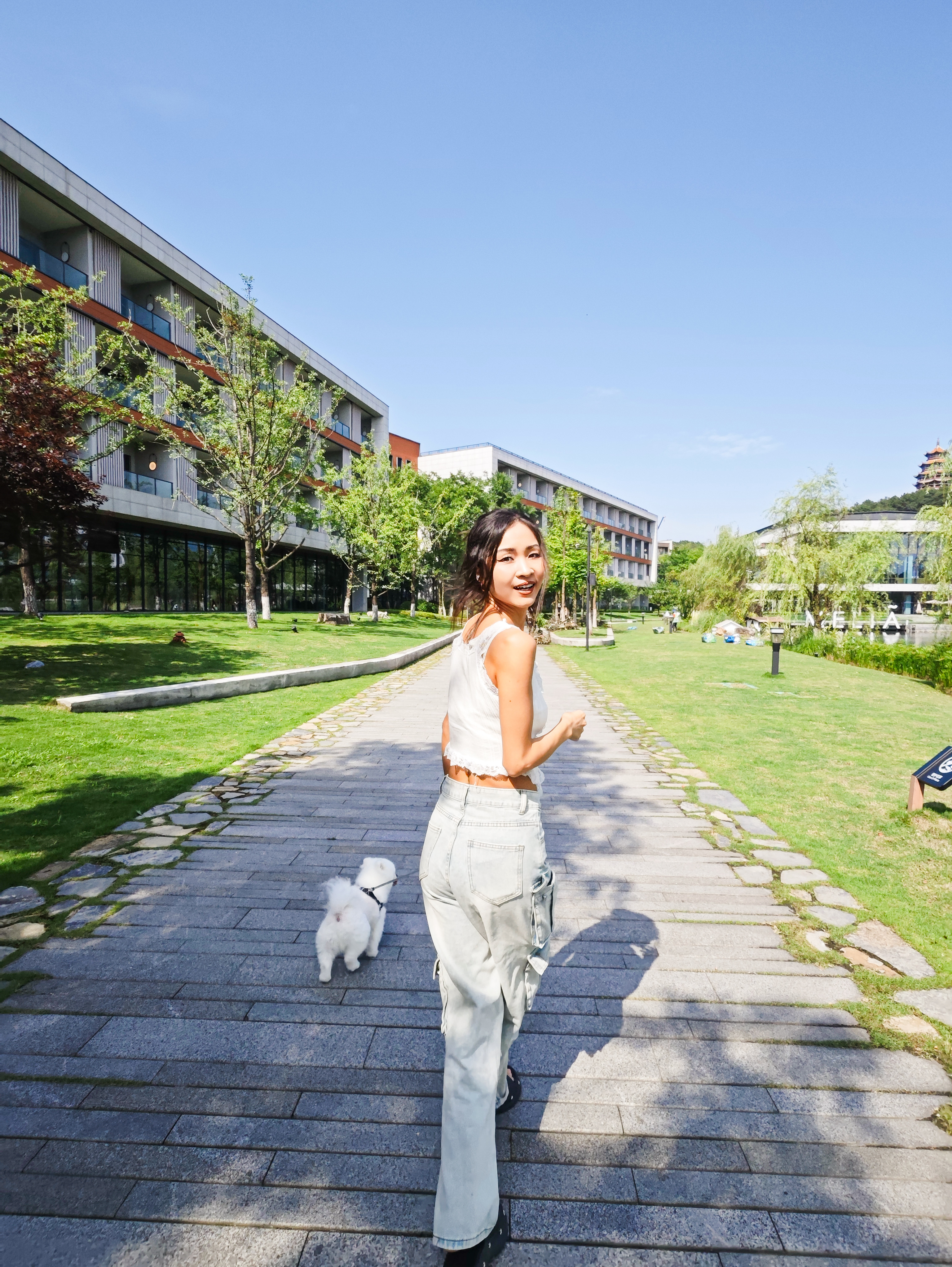 Smiling woman walks white dog on leash on stone path, modern resort buildings.