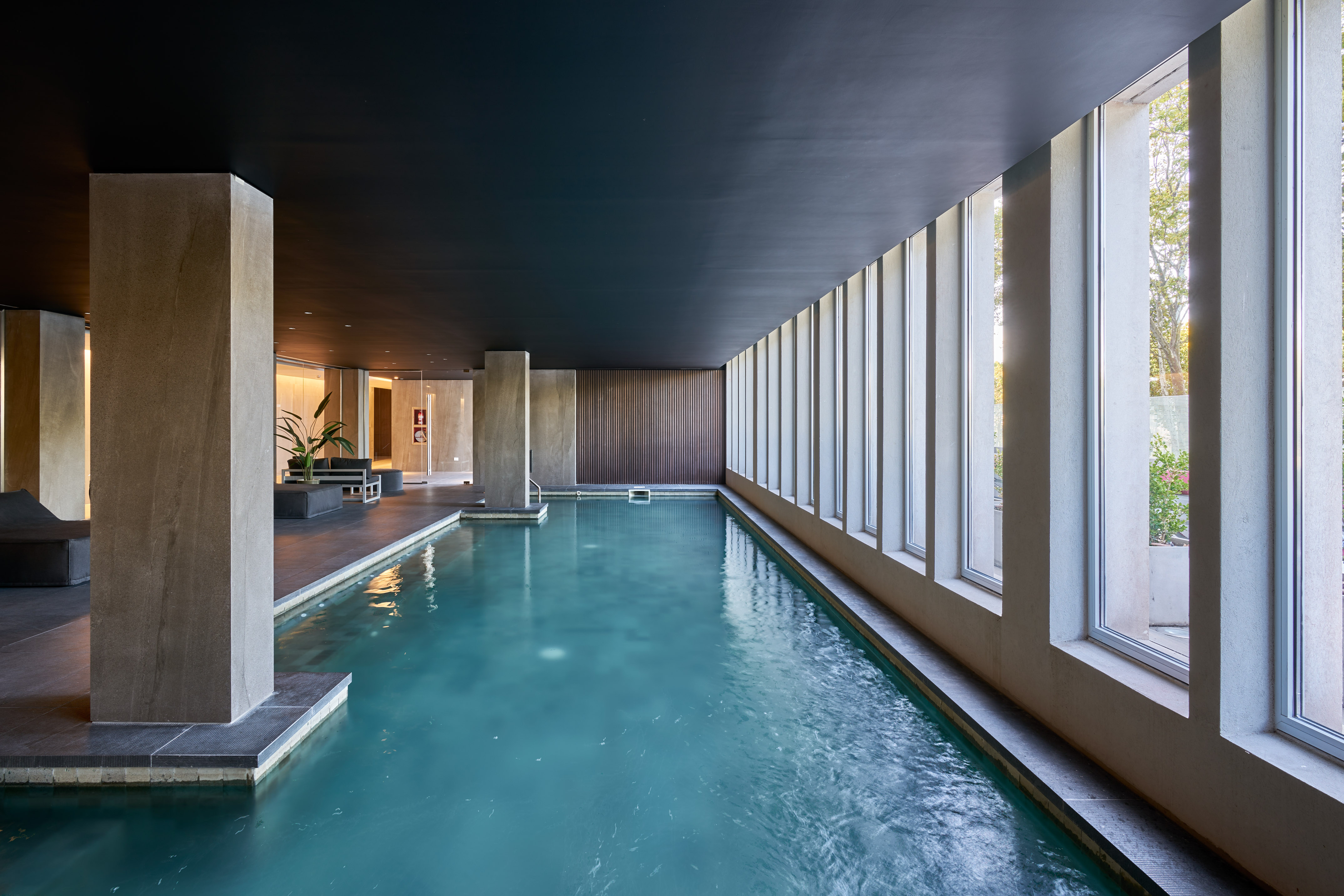 Modern indoor swimming pool with large windows, concrete columns, and dark ceiling.
