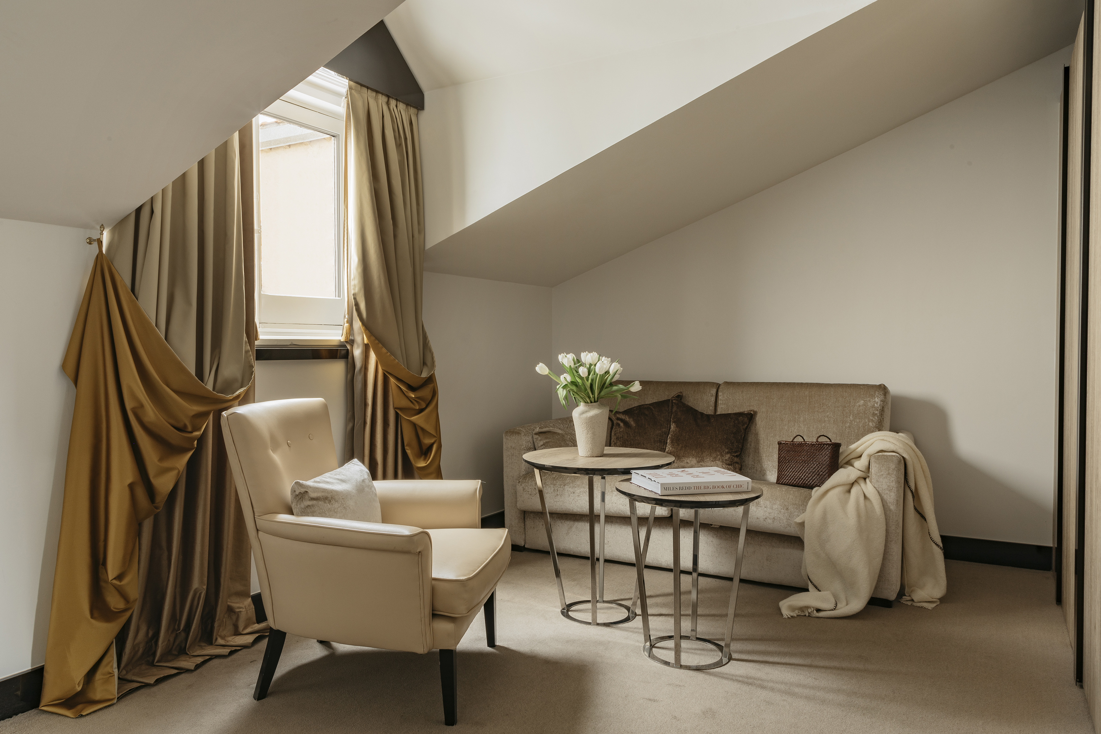 Cozy attic hotel room with slanted ceiling, neutral sofa, armchair, and window.