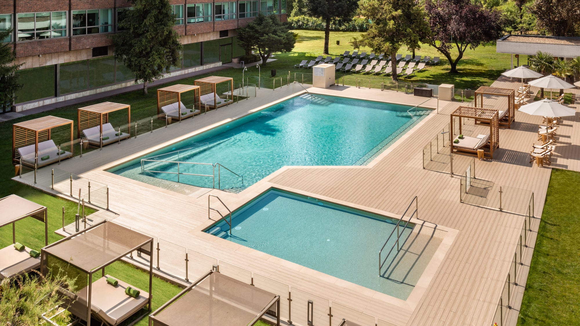 Hotel outdoor pool area with two pools, wooden deck, sunbeds, and green grass.